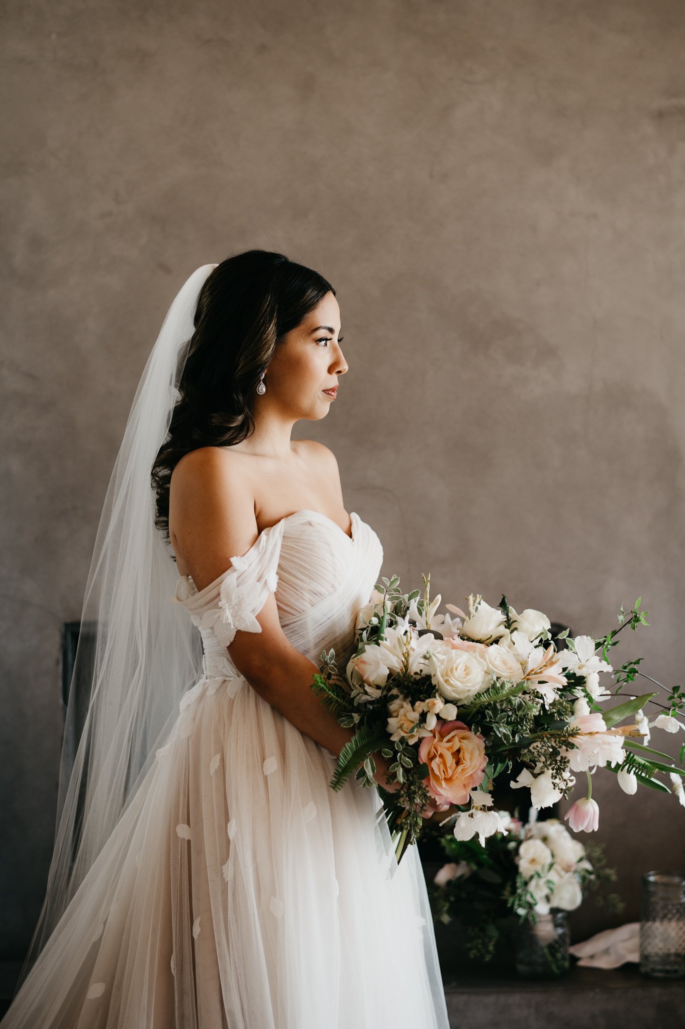 Bride in wedding dress with veil and floral bouquet staring off in distance before wedding ceremony