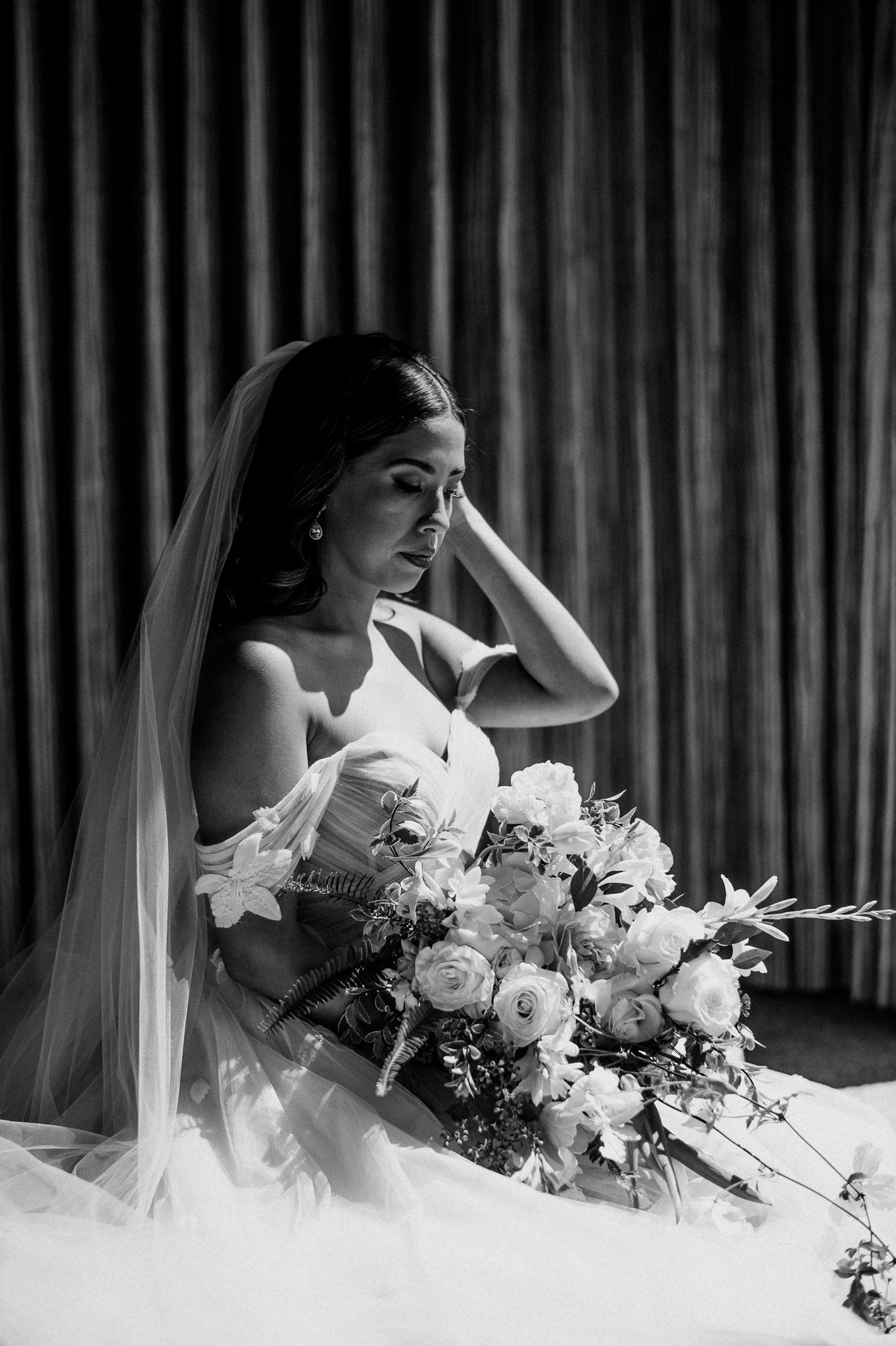 black and white photo bride in wedding dress with veil and looking at bridal bouquet before wedding ceremony