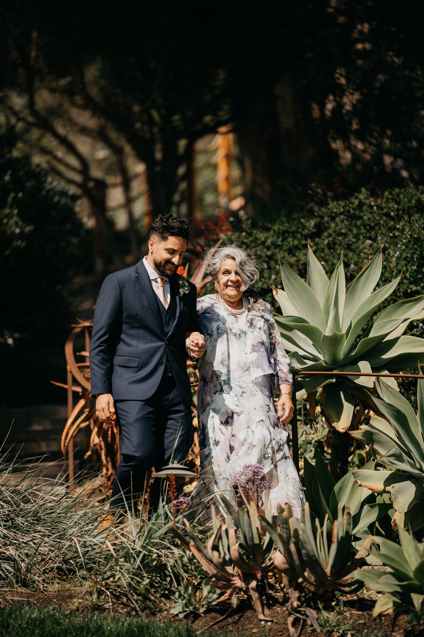 groom in blue suit walking with his mother blue and grey floral dress in garden  