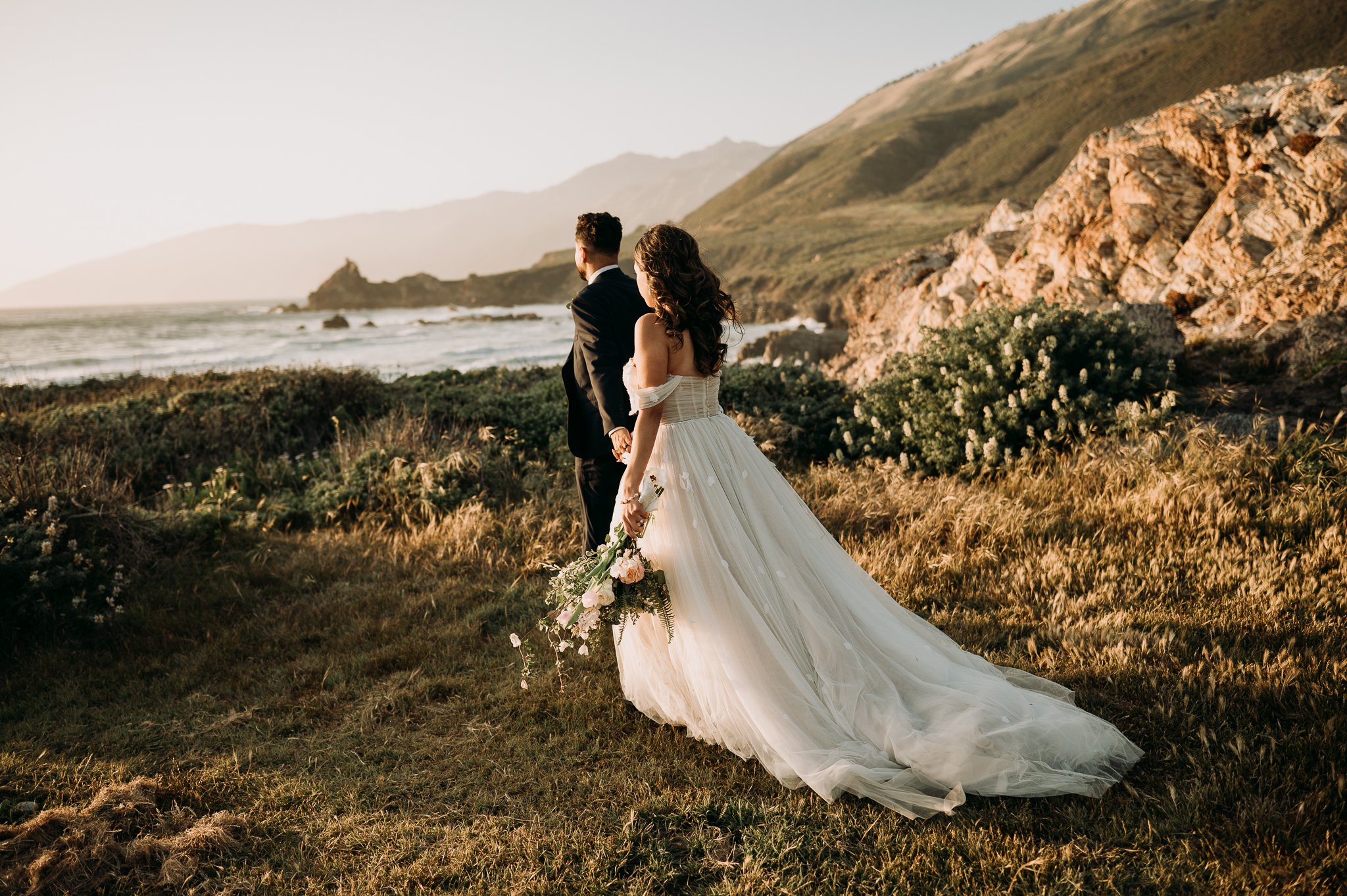 Big Sur Wedding bride and groom in wedding attire walking hand and hand toward Pacific Ocean 