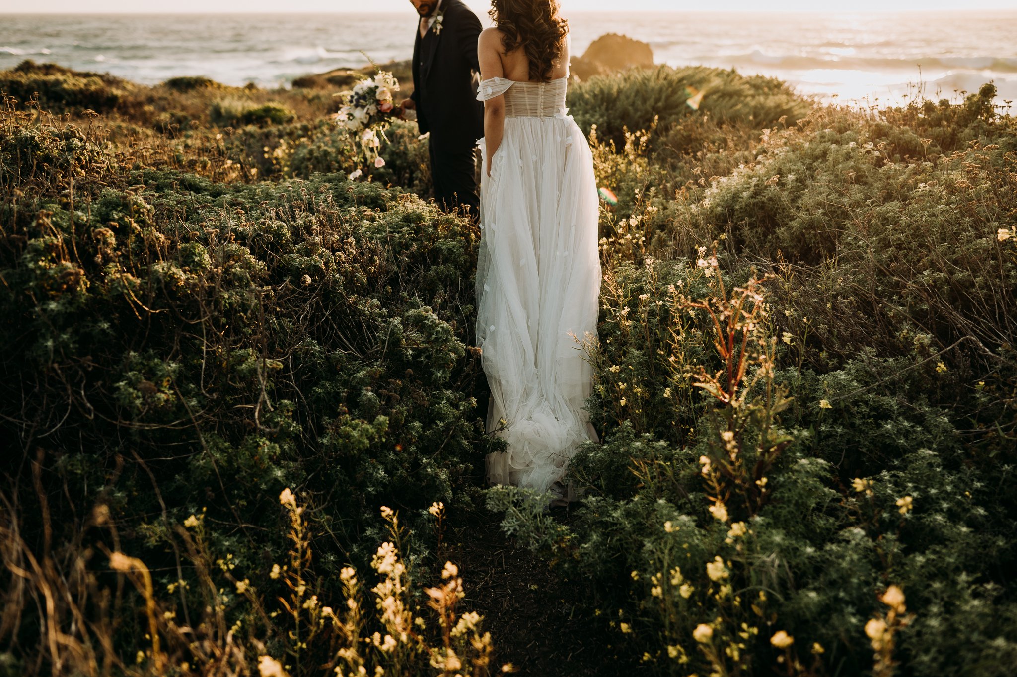 Big Sur Wedding bride and groom in wedding attire walking in field of flowers hand and hand toward Pacific Ocean
