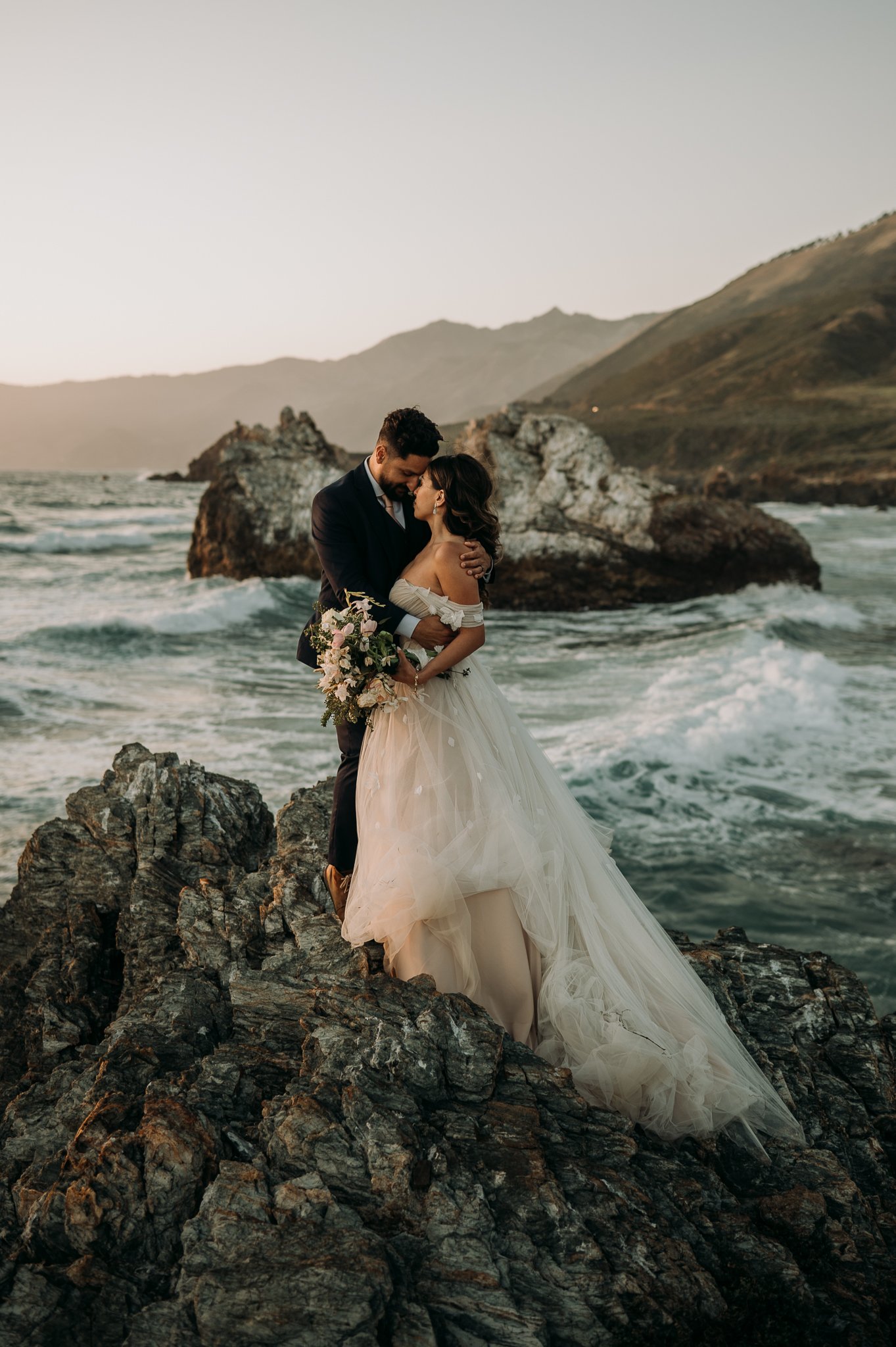 Newly married couple in wedding attire sharing a intimate moment on cliff Big Sur