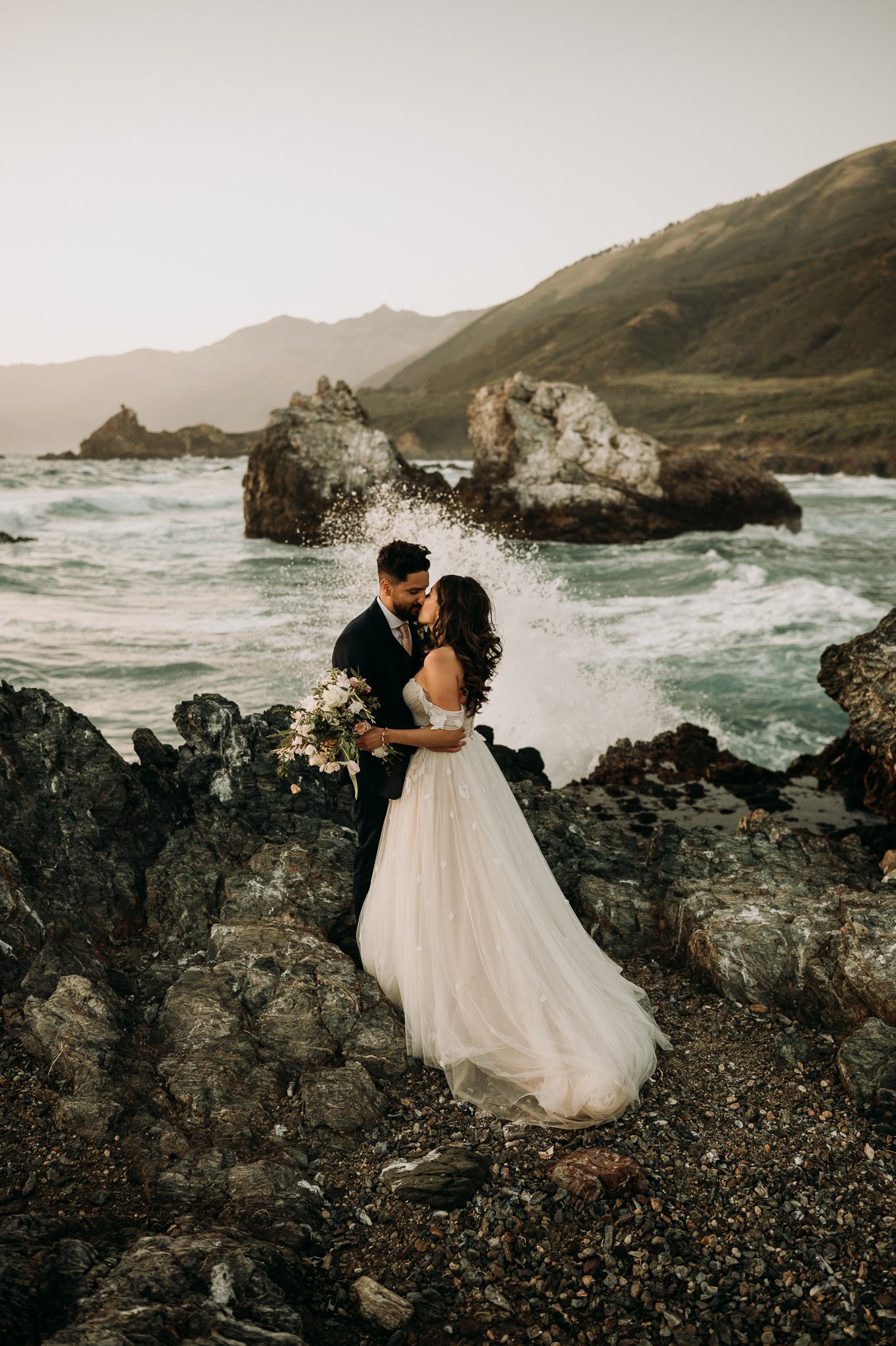 Newly married couple in wedding attire sharing a intimate moment on cliff Big Sur