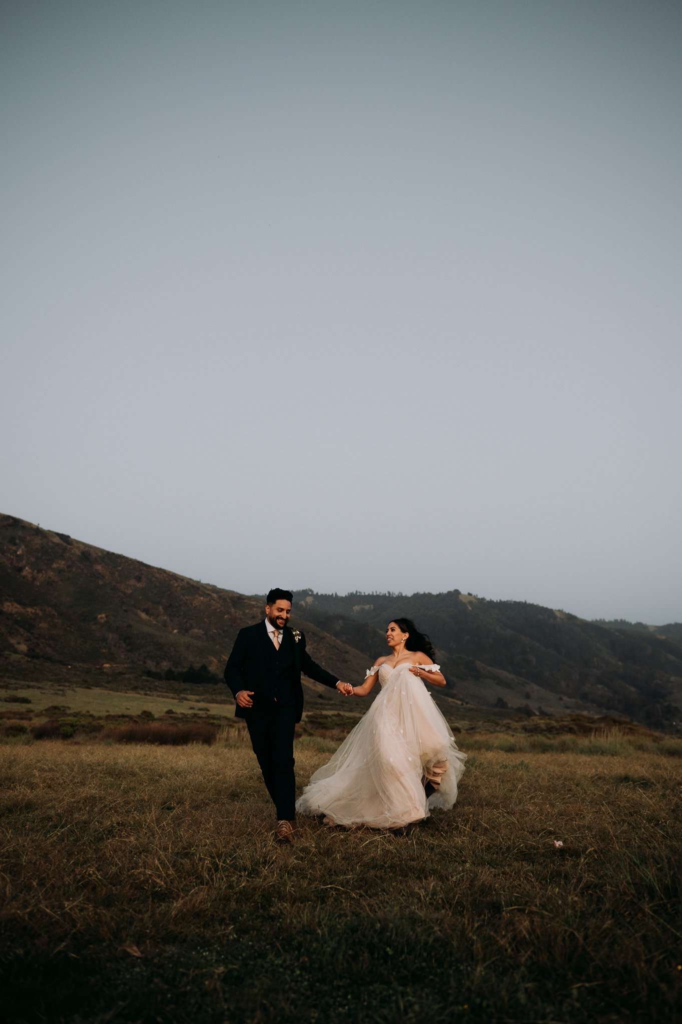 bride and groom in wedding drees and suit running hand and hand in meadow in Big Sur