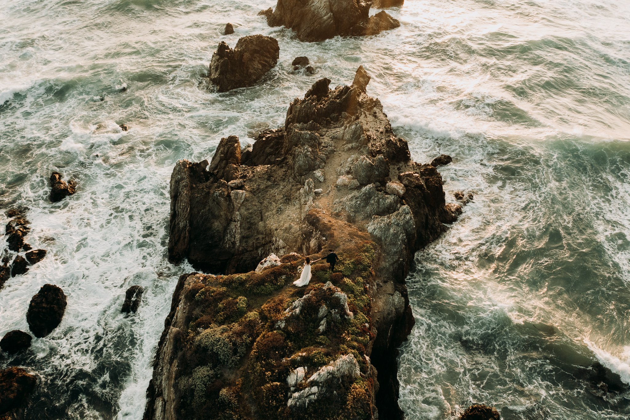 Drone photo of bride and groom walking of cliffs with Pacific ocean surrounding them both