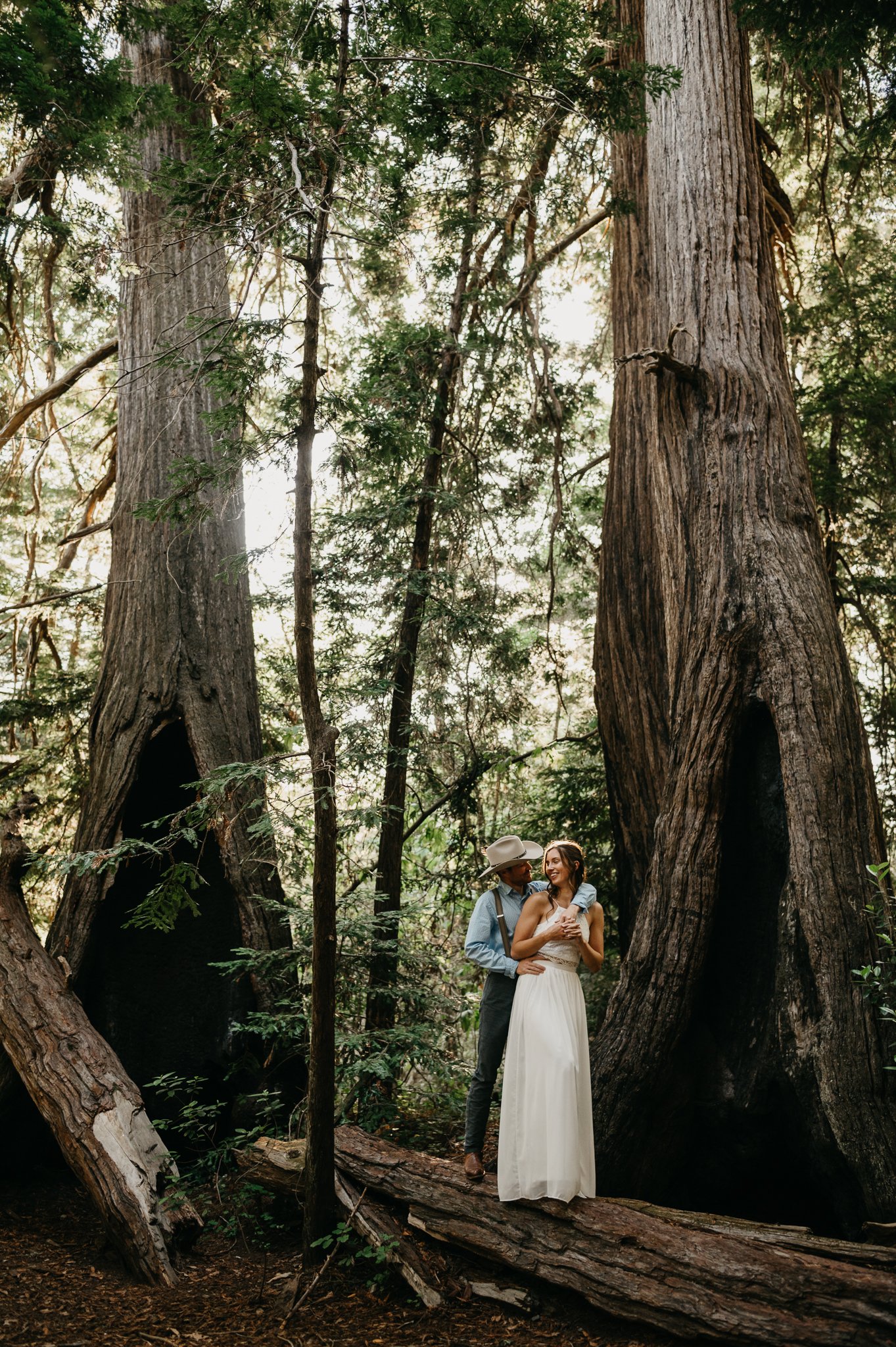 Big Sur Engagement couple in forest kissing under redwoods she in long white sleeveless dress and he is in a light blue shit and tan pants and cowboy hat