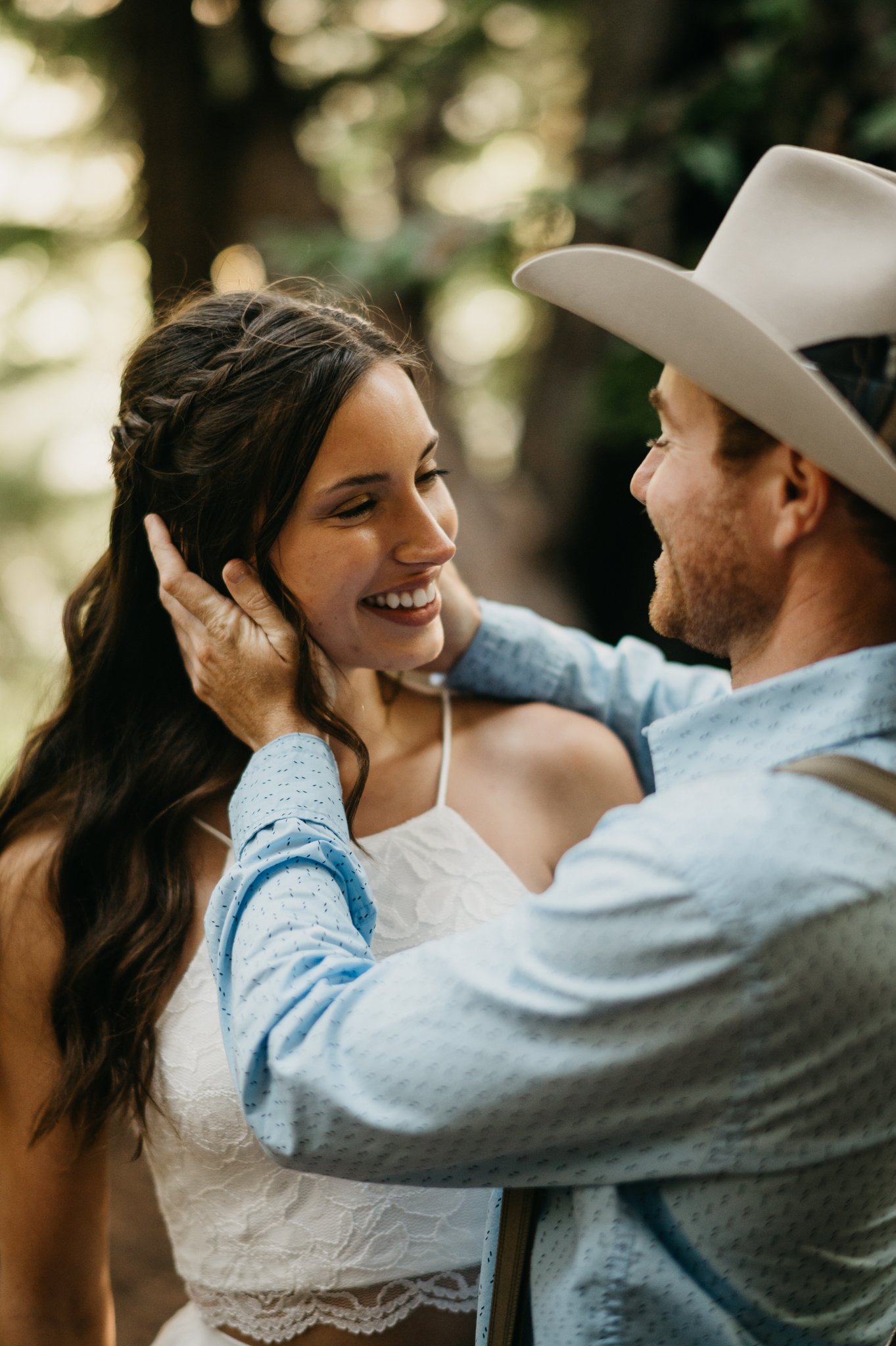 Big Sur Engagement couple in forest embracing under redwoods she in long white sleeveless dress and he is in a light blue shit and tan pants and cowboy hat