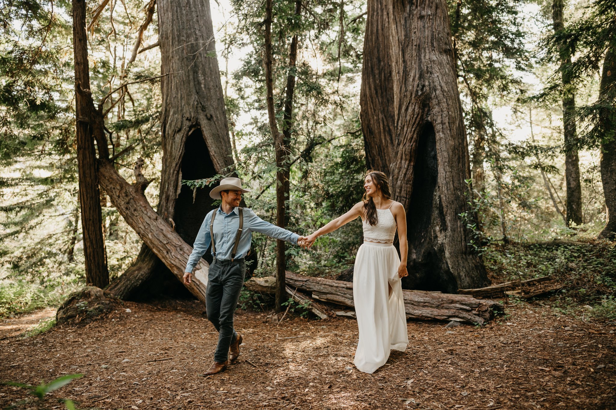 Big Sur Engagement couple in forest holding hands walking under redwoods she in long white sleeveless dress and he is in a light blue shit and tan pants and cowboy hat