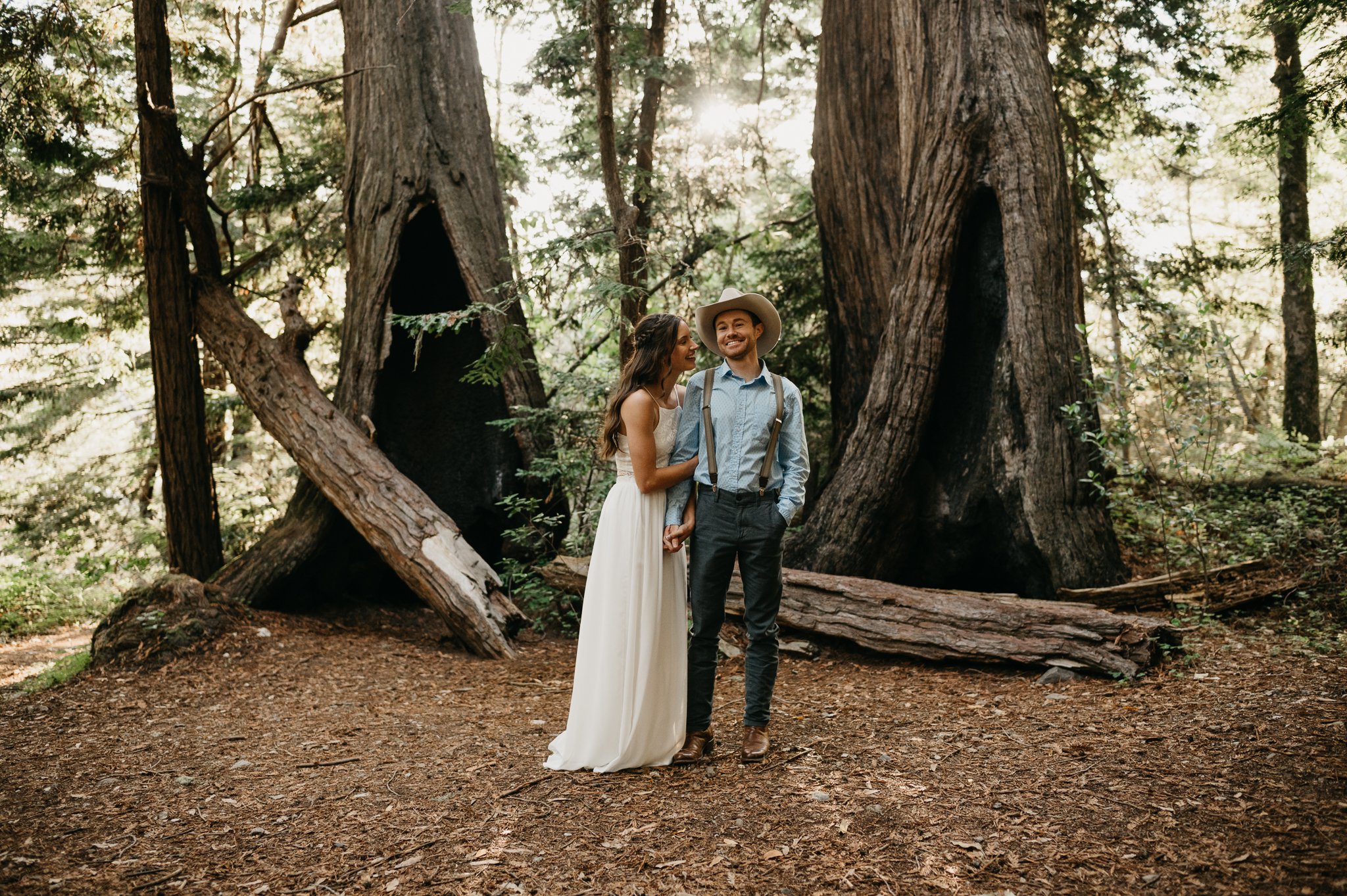Big Sur Engagement couple in forest hugging under redwoods she in long white sleeveless dress and he is in a light blue shit and tan pants and cowboy hat