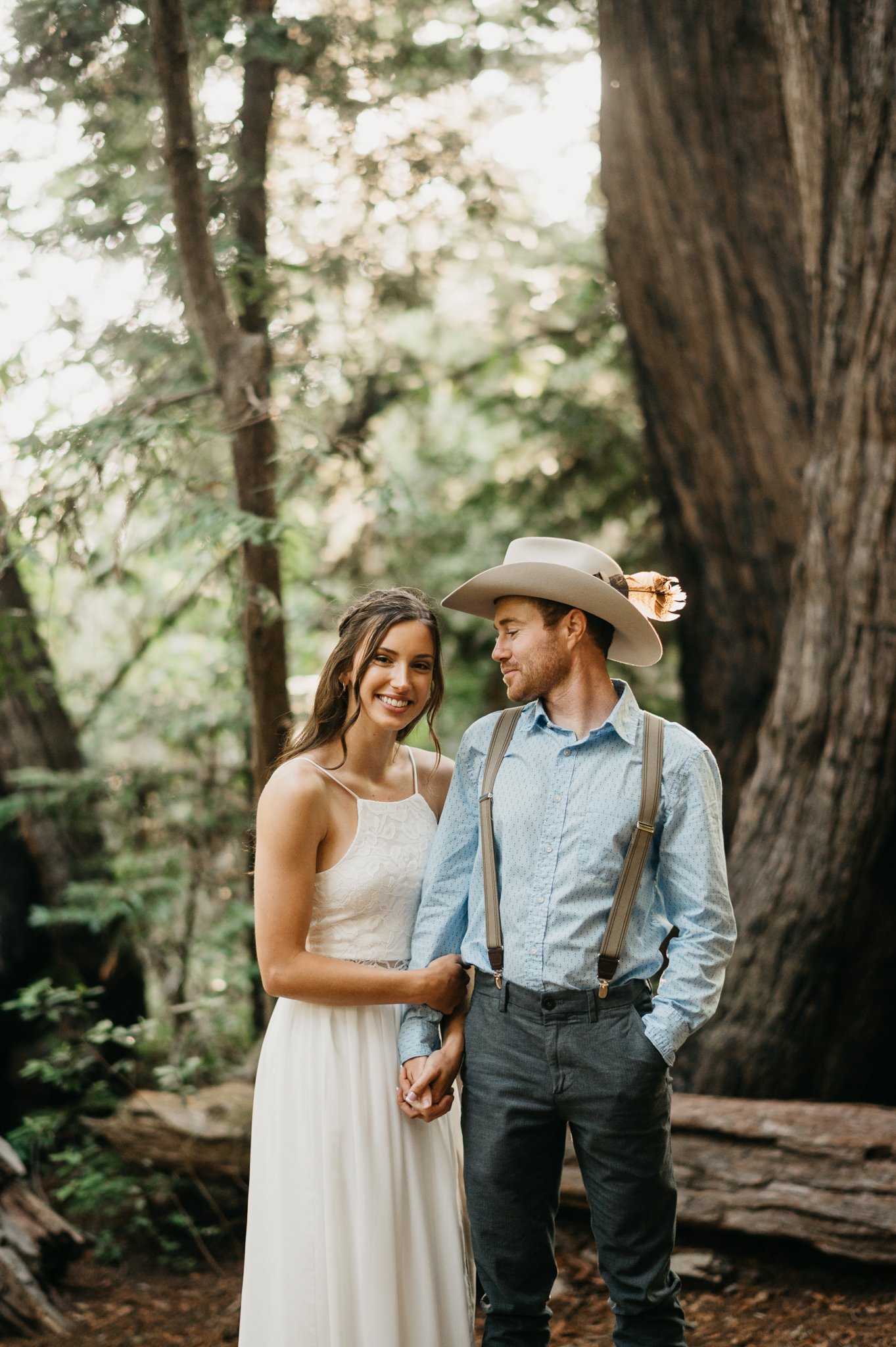 Big Sur Engagement couple in forest embracing under redwoods she in long white sleeveless dress and he is in a light blue shit and tan pants and cowboy hat