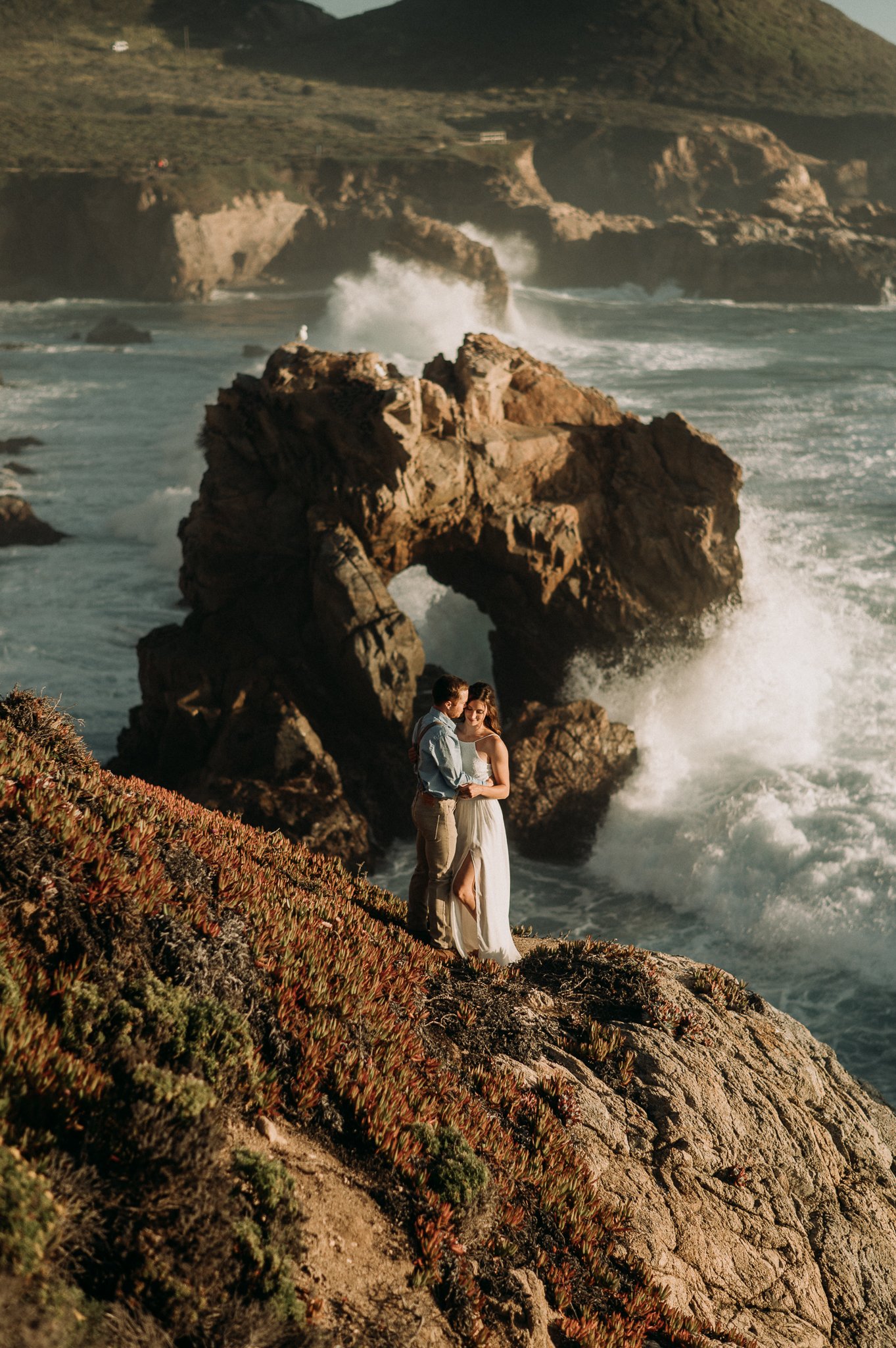 Couple cliffside at keyhole rock in Big Sur couple hugging looking out at Pacific Ocean 