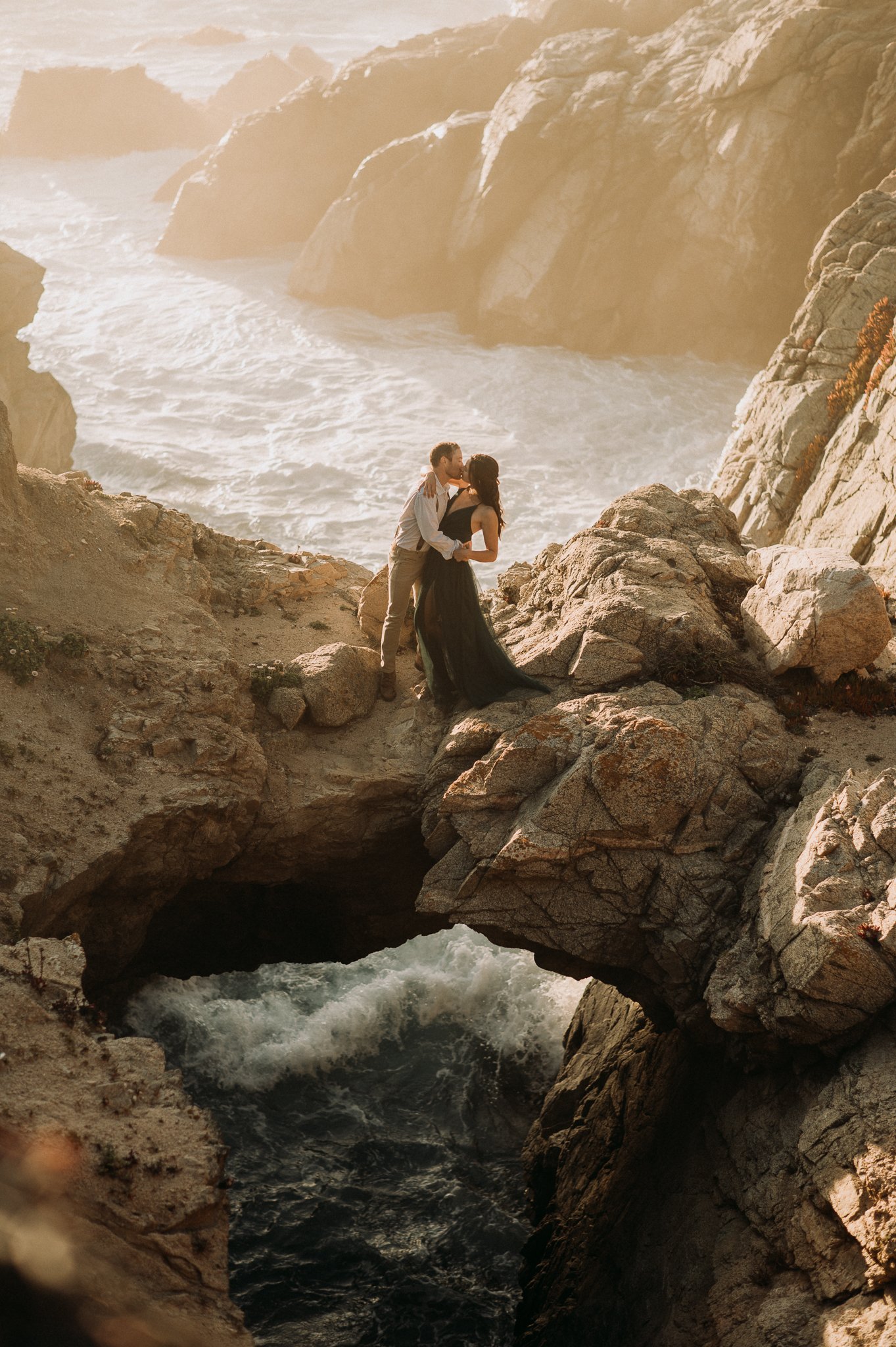 Newly engaged couple hugging at Big Sur, California on cliffs with Pacific Ocean waves behind them