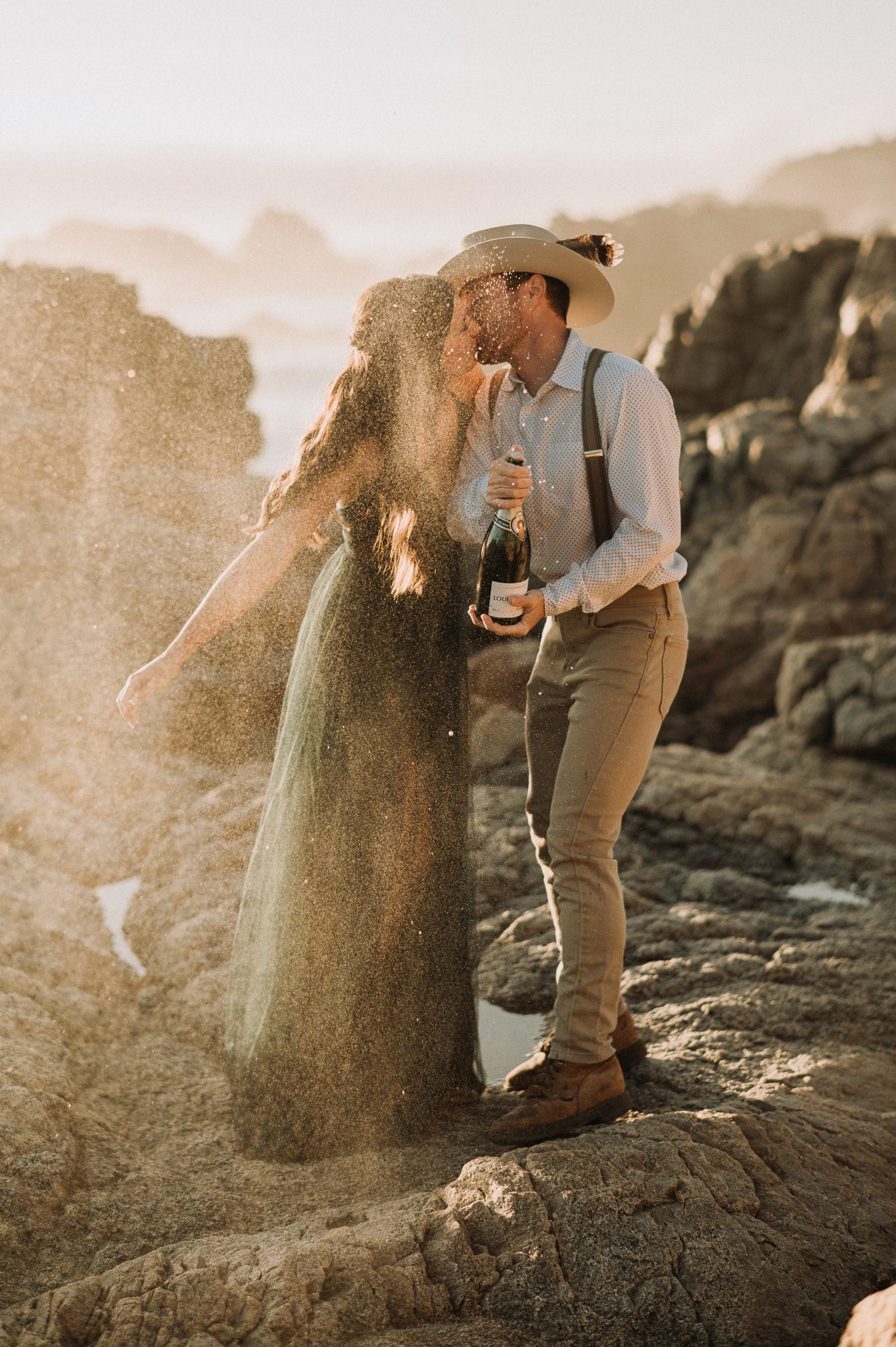 Newly engaged couple kissing popping champagne cliffside 
