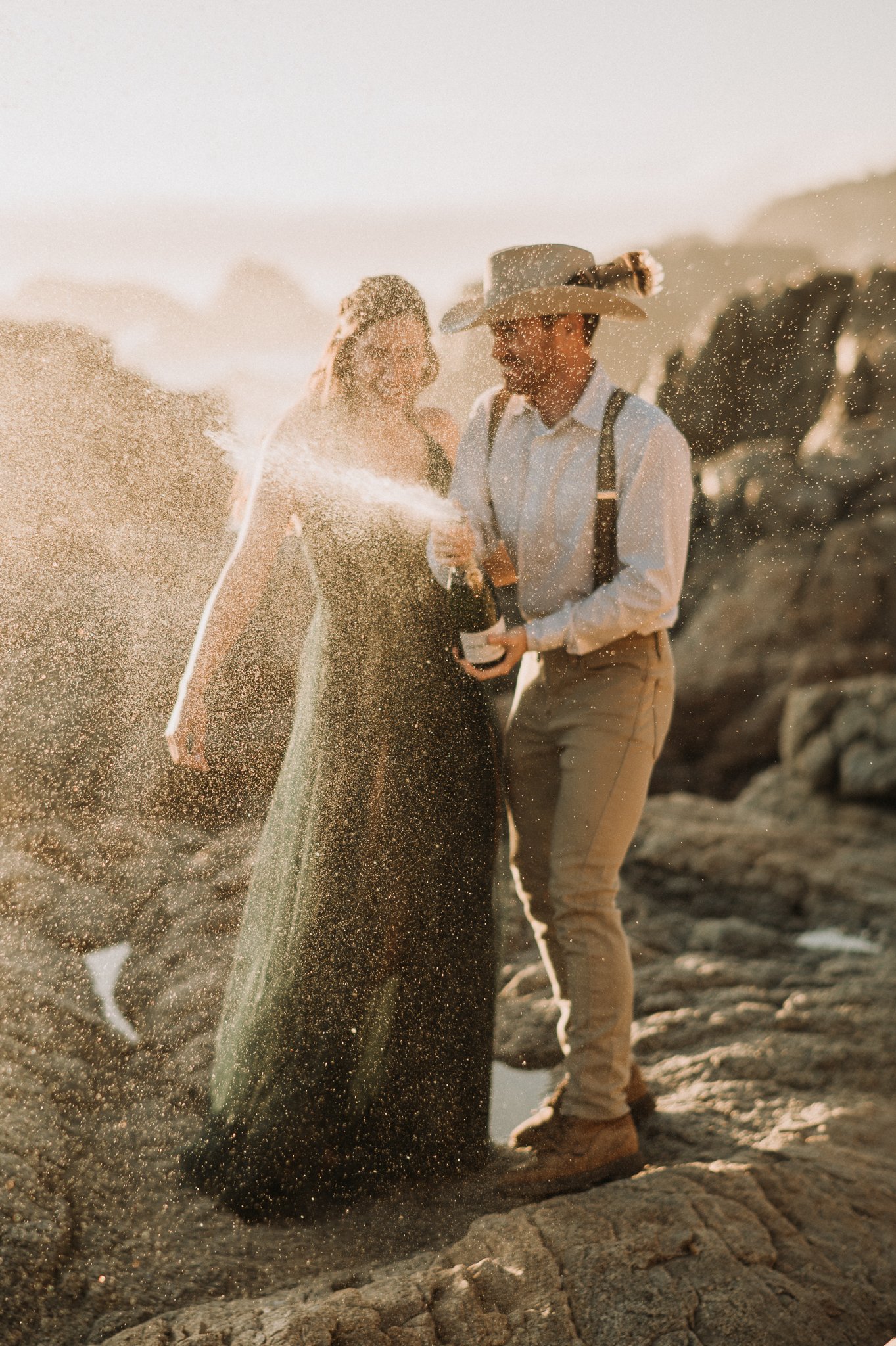 Newly engaged couple at Big Sur, California on cliffs popping champagne at sunset 