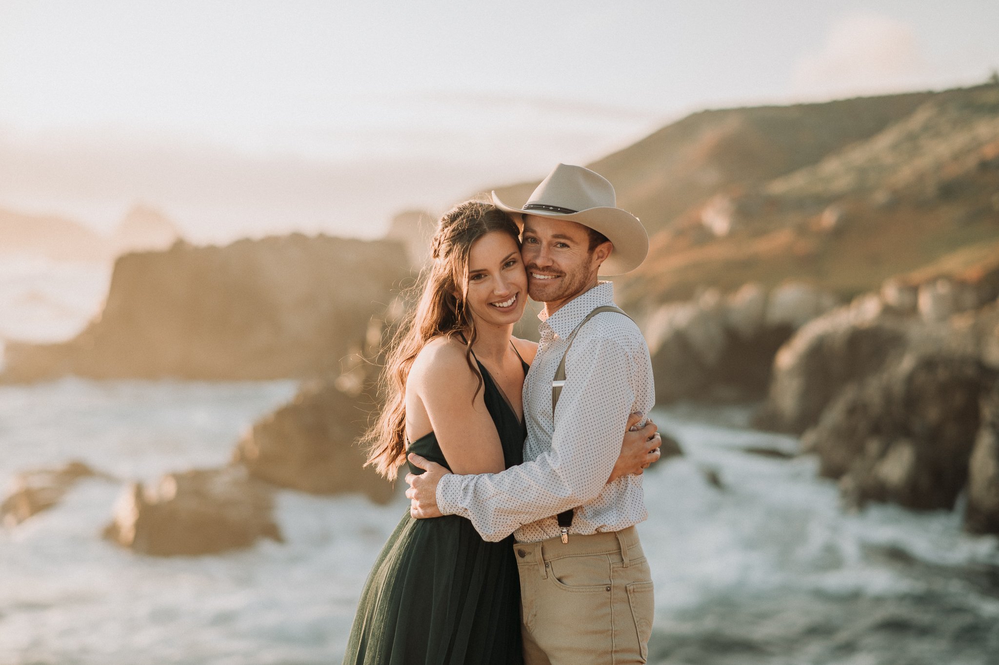 Newly engaged couple posing on cliff with Pacific ocean in background 