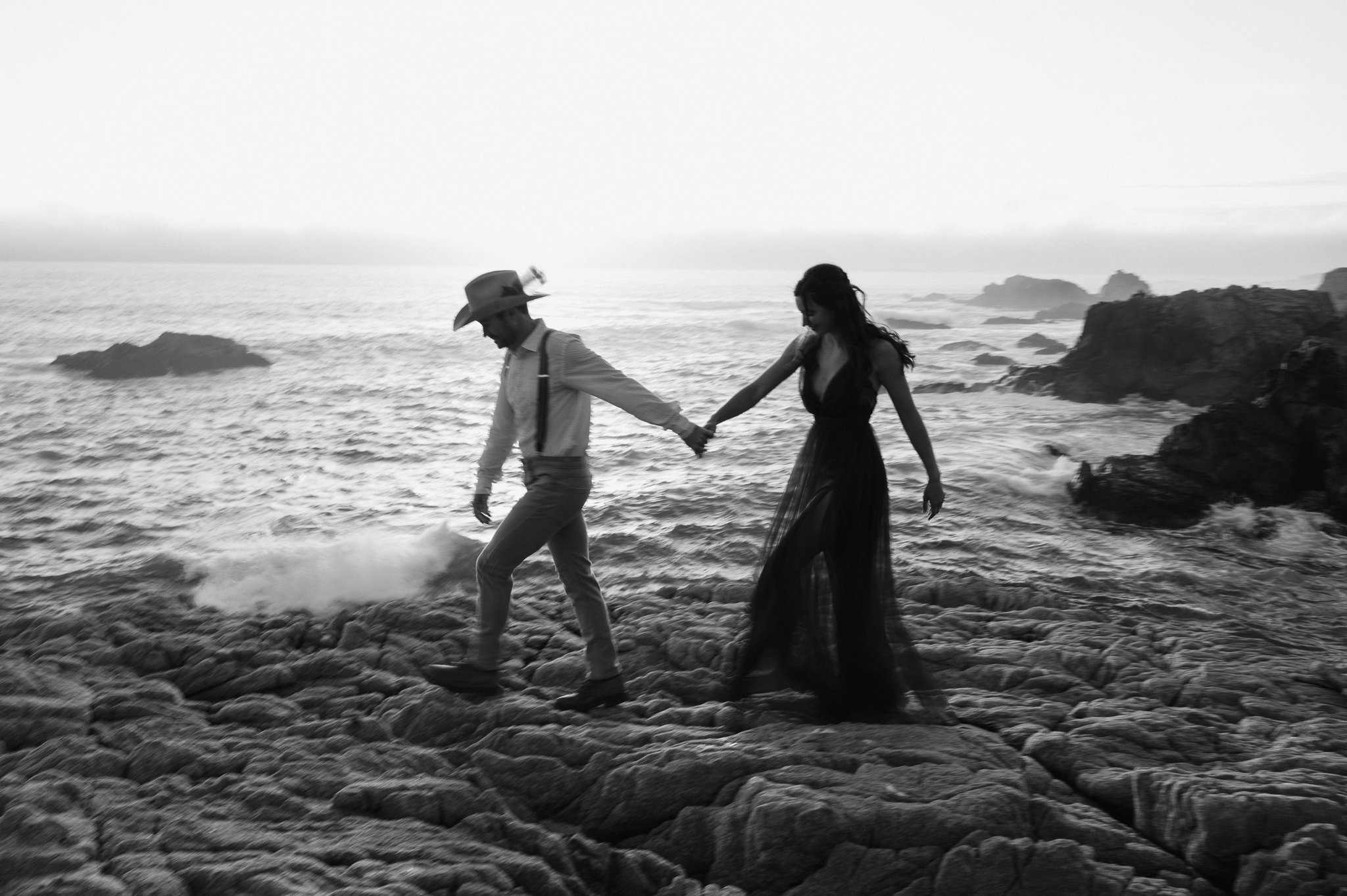 Black and white photo newly engaged couple walking along cliffs with Pacific ocean in background 