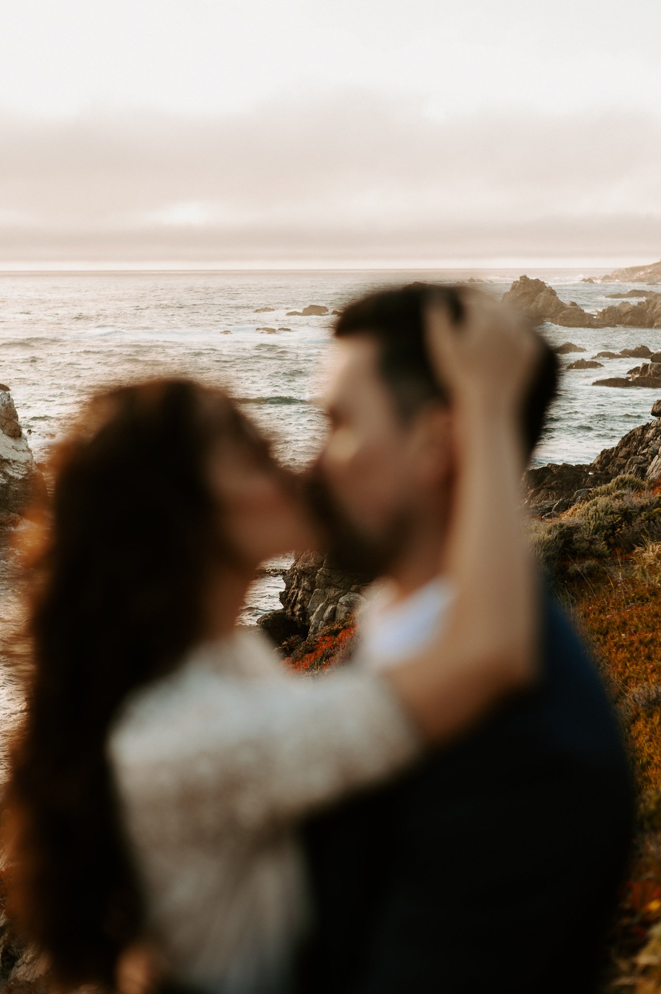 Newly engaged couple upper bodies showing kissing on cliff above Pacific Ocean in background