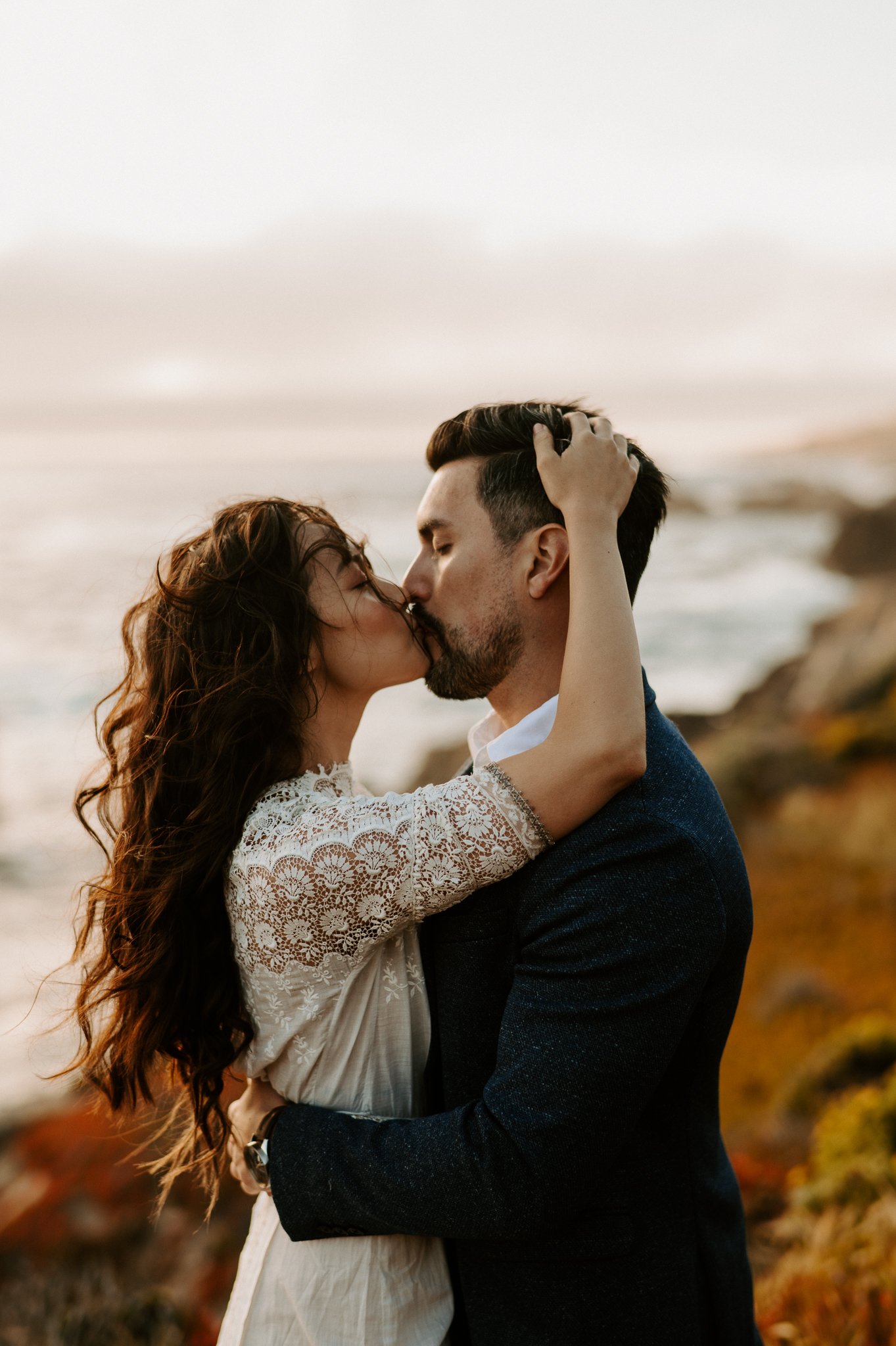 Newly engaged couple upper bodies showing kissing on cliff above Pacific Ocean in background