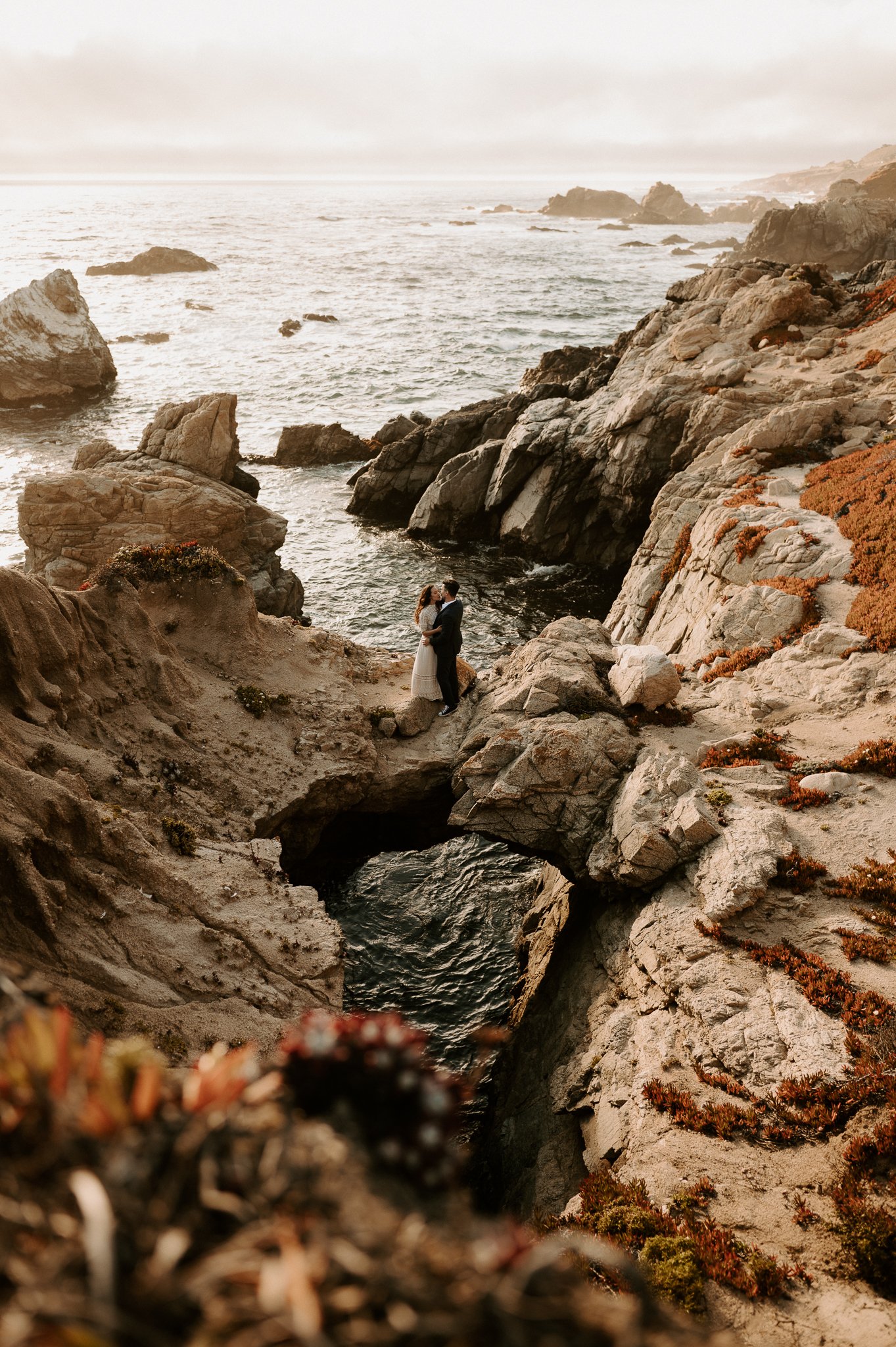 couple on large rock holding each other intimately with the ocean waves under the key hole in rock Big Sur, California