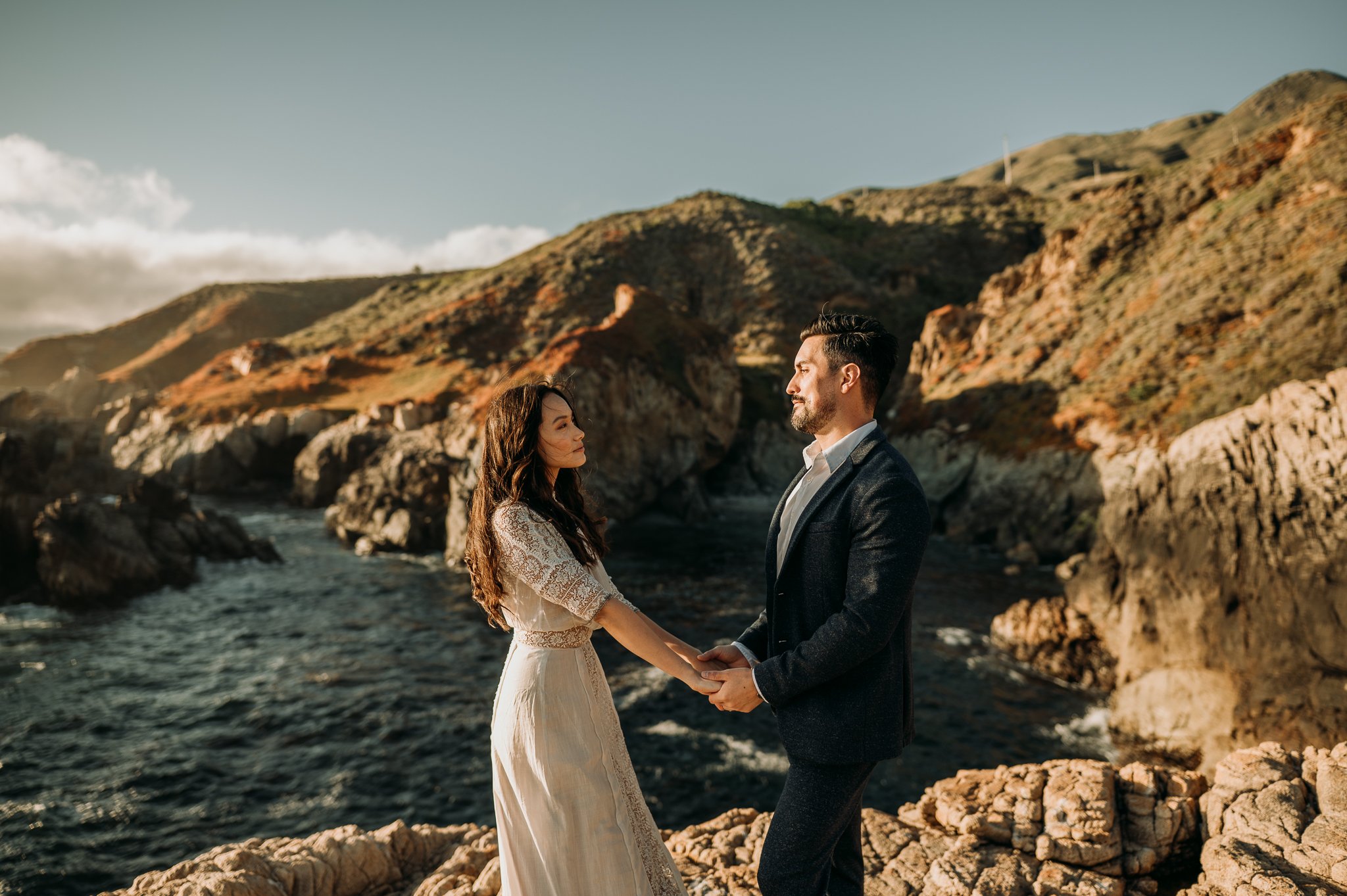 couple cliffside facing each other holding hands with ocean in background in Big Sur
