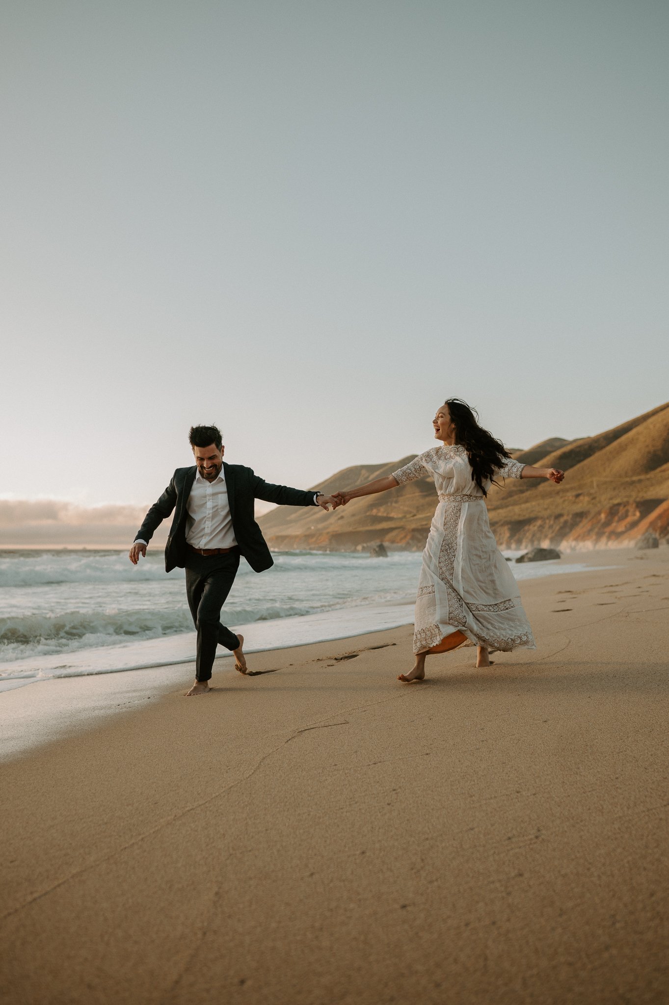 Newly engaged couple holding hand running on beach in Big Sur