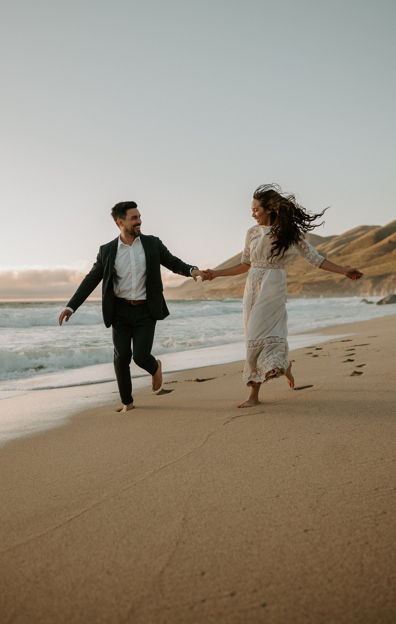 Newly engaged couple holding hand running on beach in Big Sur