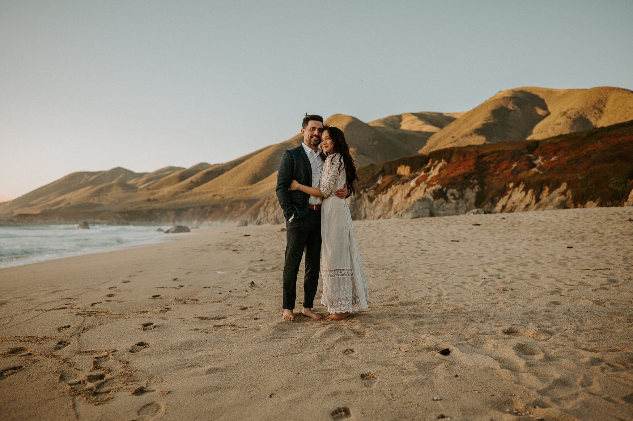Newly engaged couple hugging looking at camera on beach Big Sur