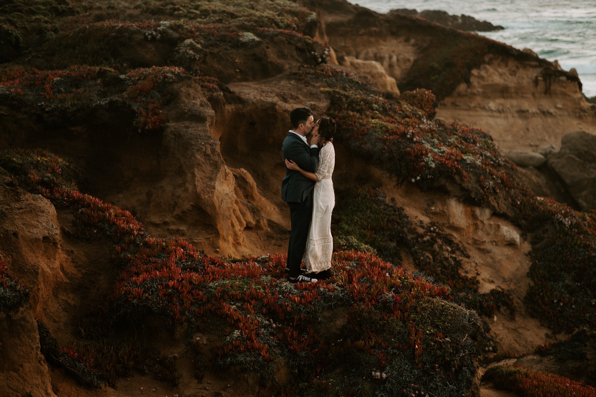 newly engaged couple standing on cliff in Big Sur opening champagne sharing a kiss