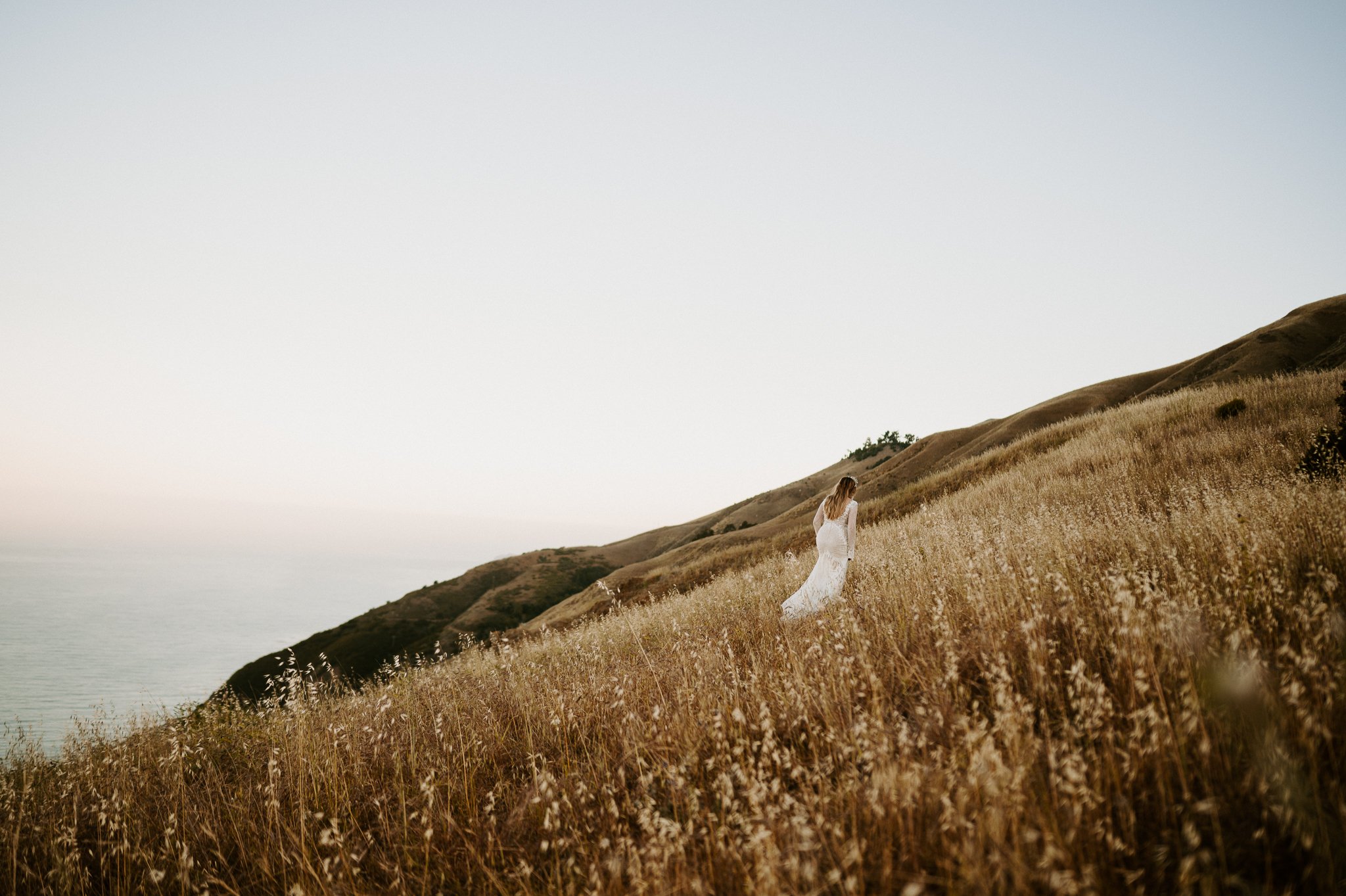 Bride in wedding dress walking up hill in tall brown grasses in Big Sur