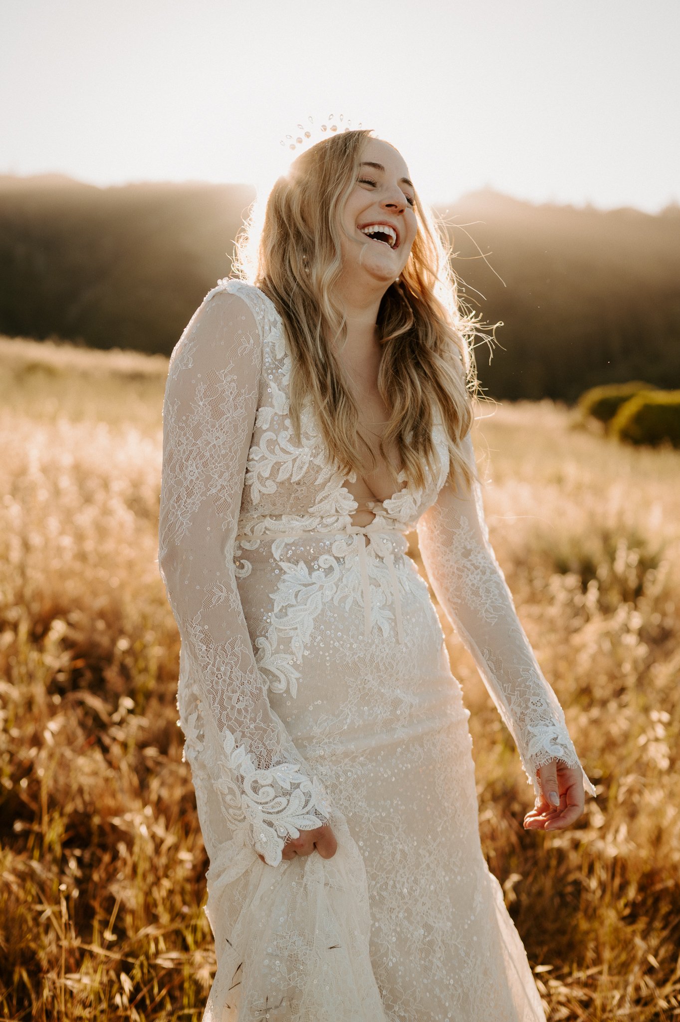 Big Sur Bride posing in tall grass, in lace wedding dress she is laughing