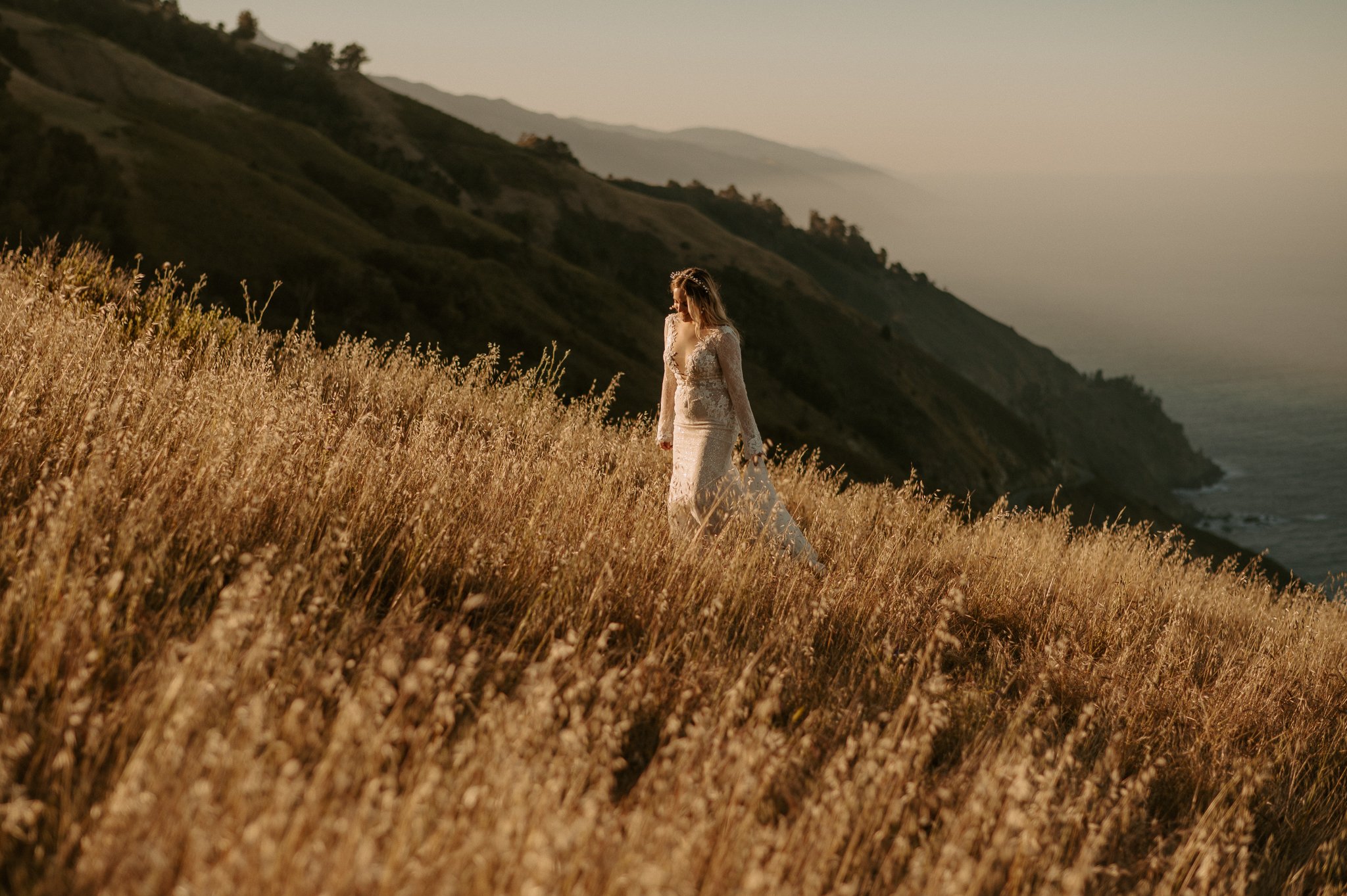 Big Sur Bride walking uphill in tall grass, in lace wedding dress.
