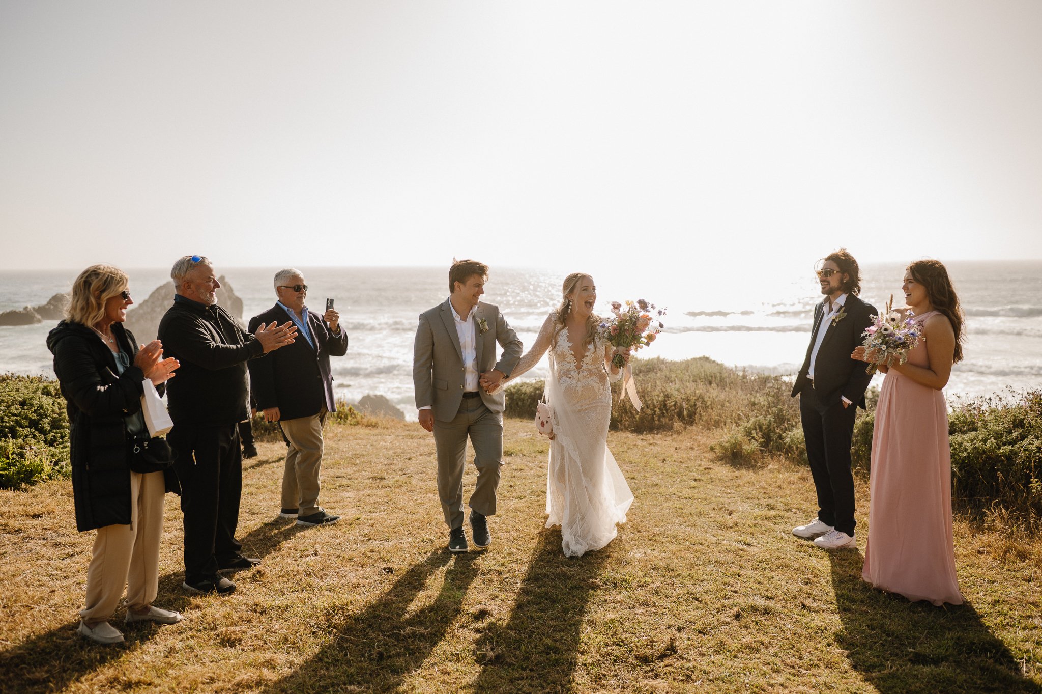 bride and groom walking toward camera surrounded by family on grassy cliff in Big Sur