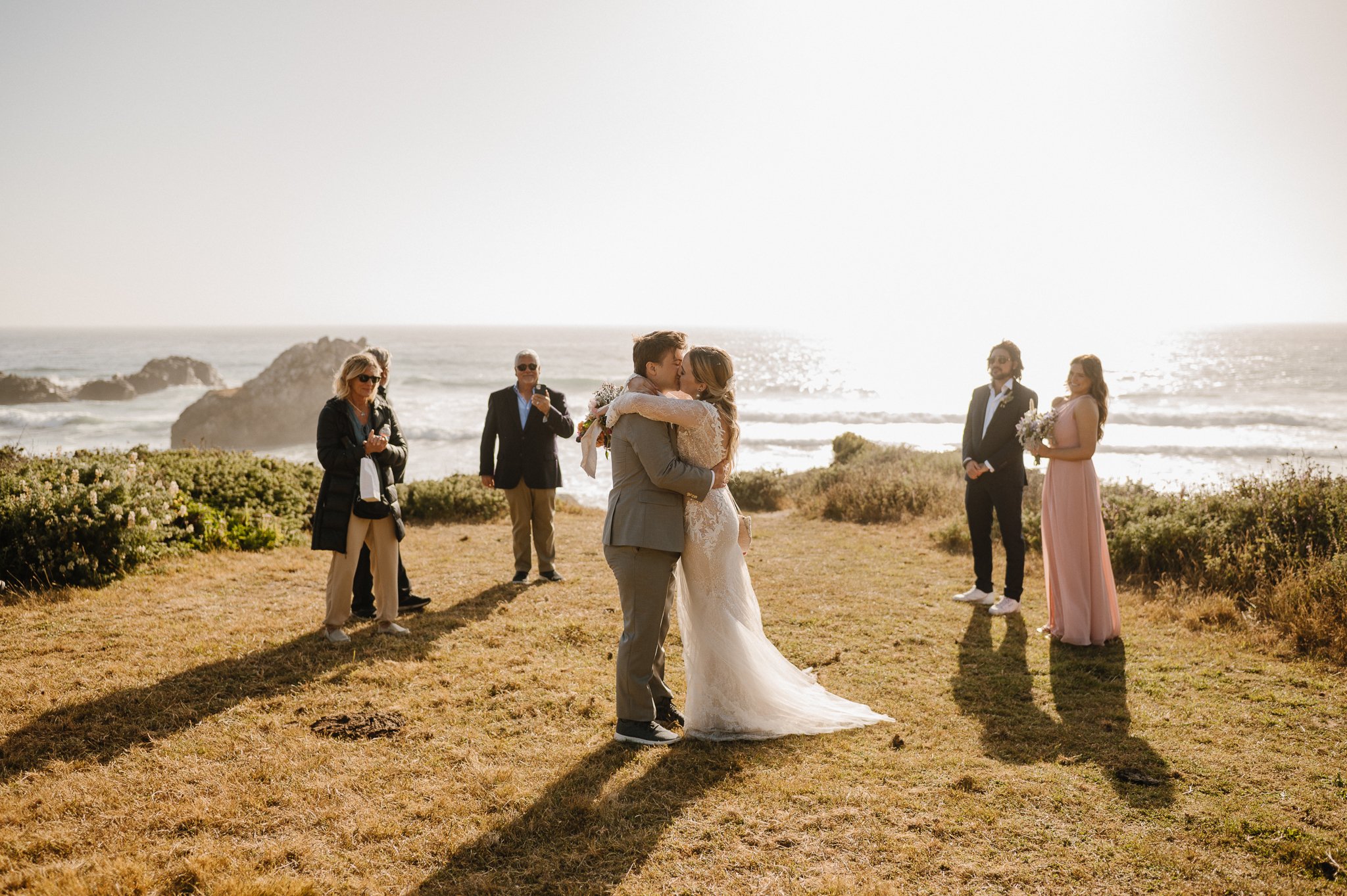 bride and groom kissing after ceremony surrounded by family on grassy cliff in Big Su