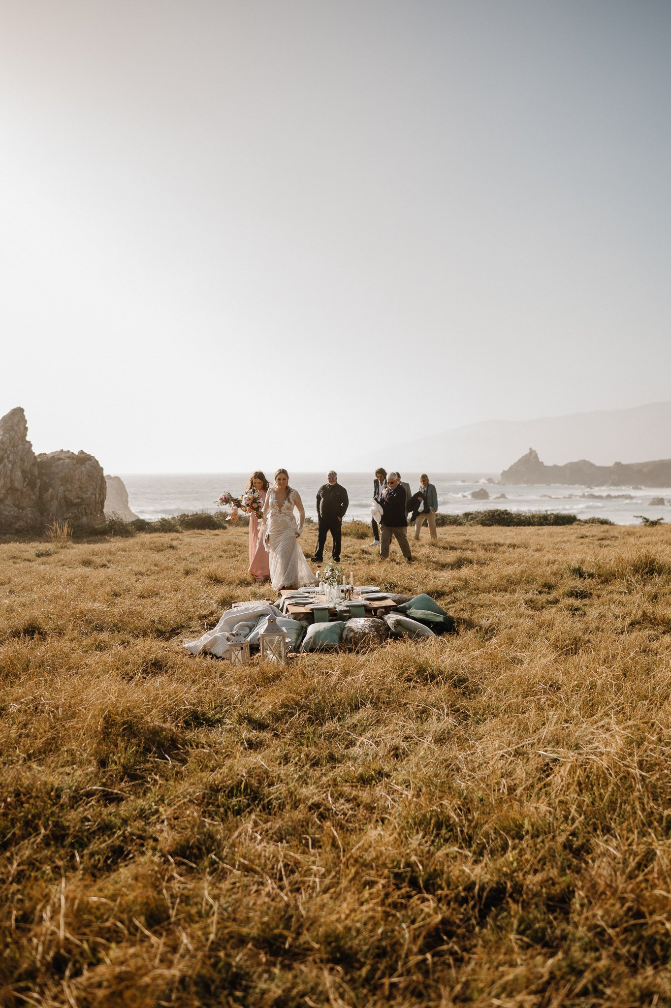 Big Sur Wedding cliffside picnic, table setup in grass with plates, glassware and pillows for all bride, groom and family