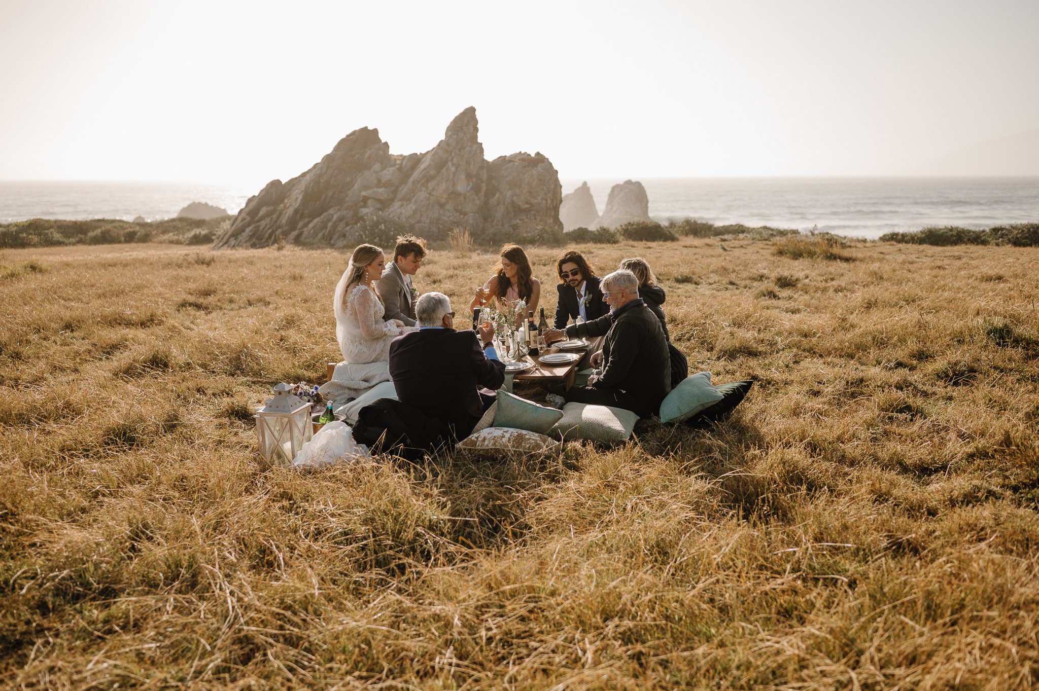 Big Sur Wedding picnic on cliff with bride, groom and family sitting around table, Pacific Ocean in background.