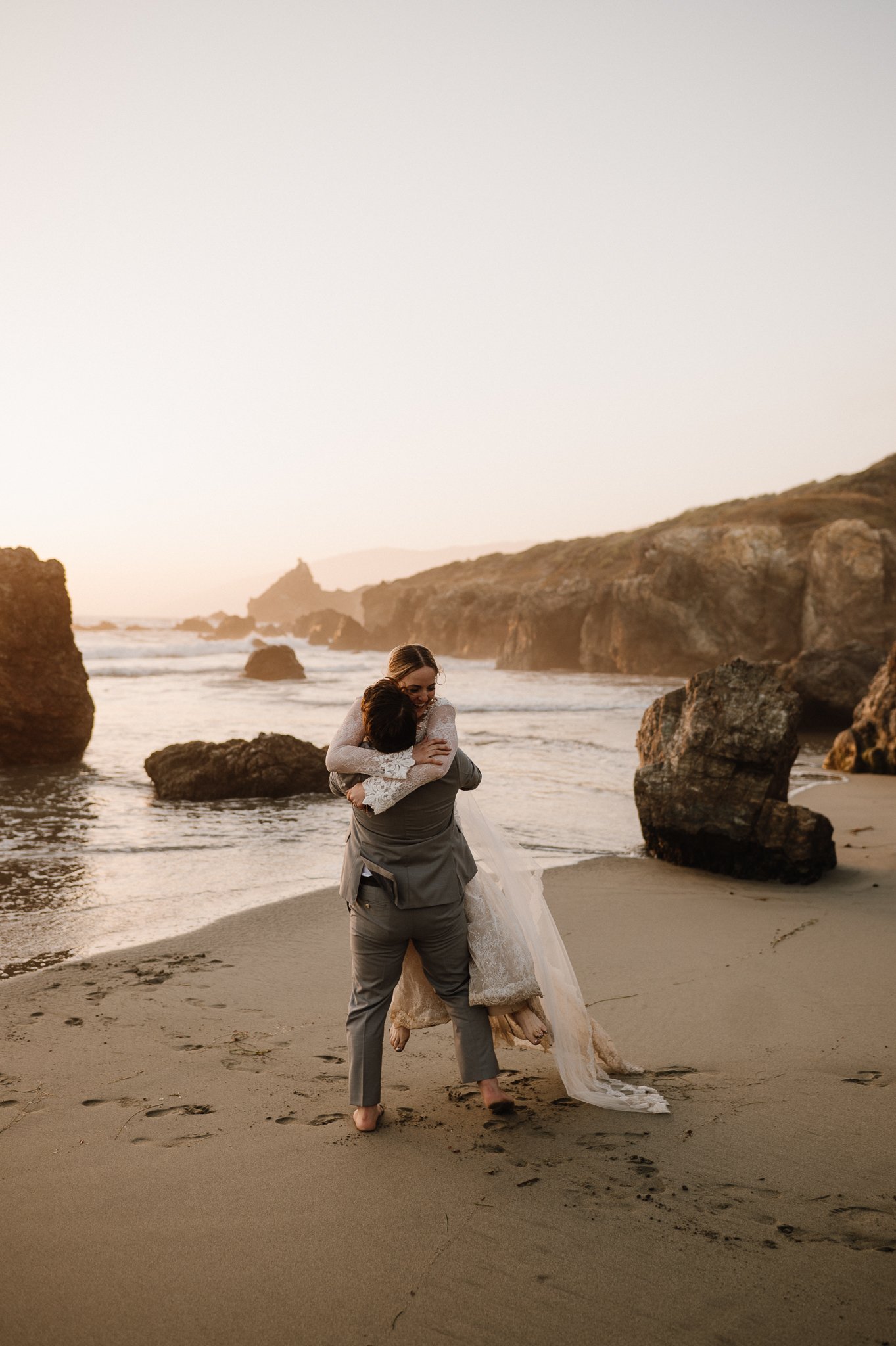 Big Sur bride and groom on beach, groom in lifting bride off the ground hugging her, bride is smiling