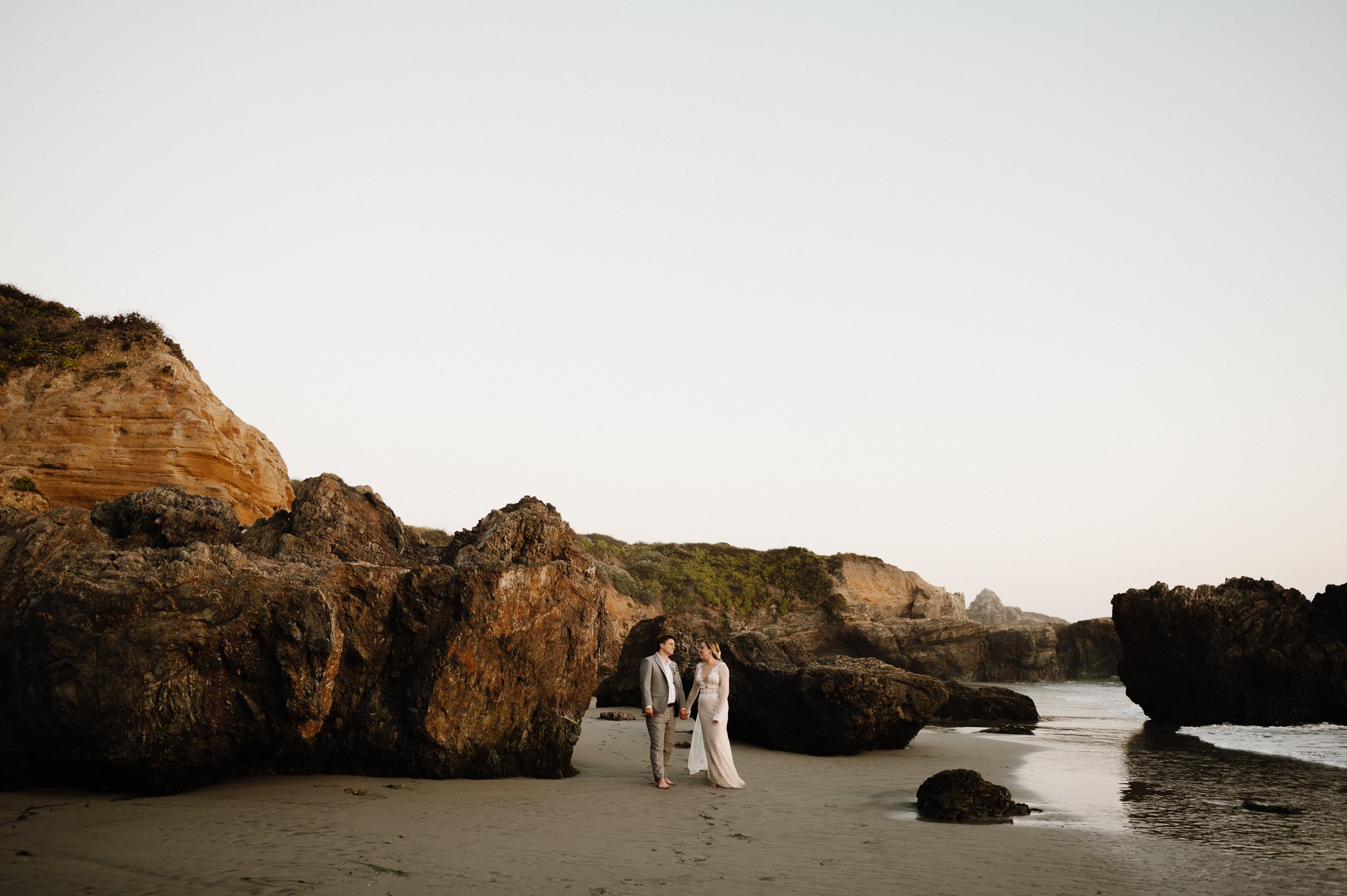 Big Sur bride and groom on beach walking in sand with cliffs and hills in background looking and each other smiling