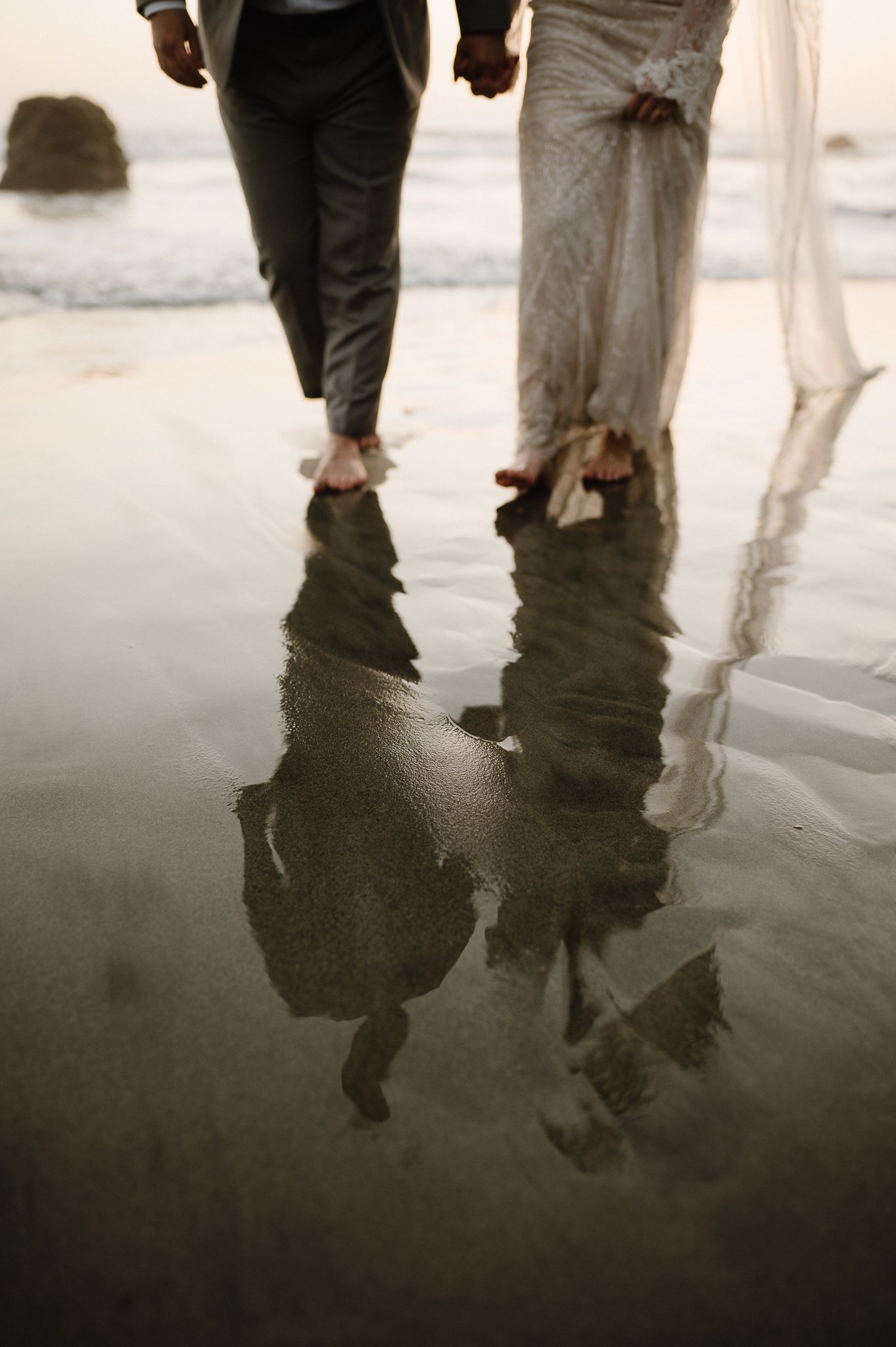 Bride and grooms legs on sandy beach with reflection of themselves in wet sand