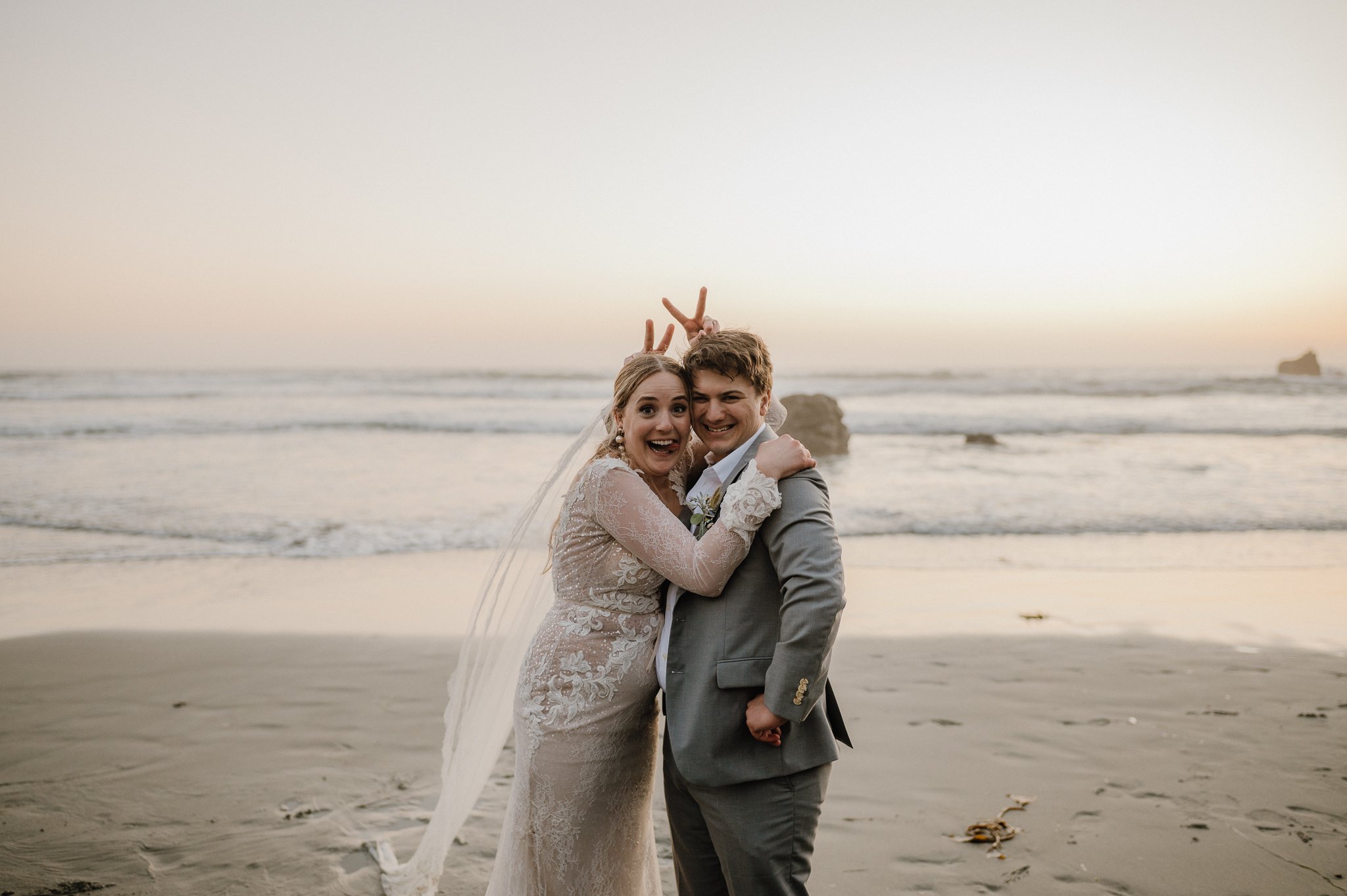 bride and groom on beach making funny faces making bunny ears behind each others heads