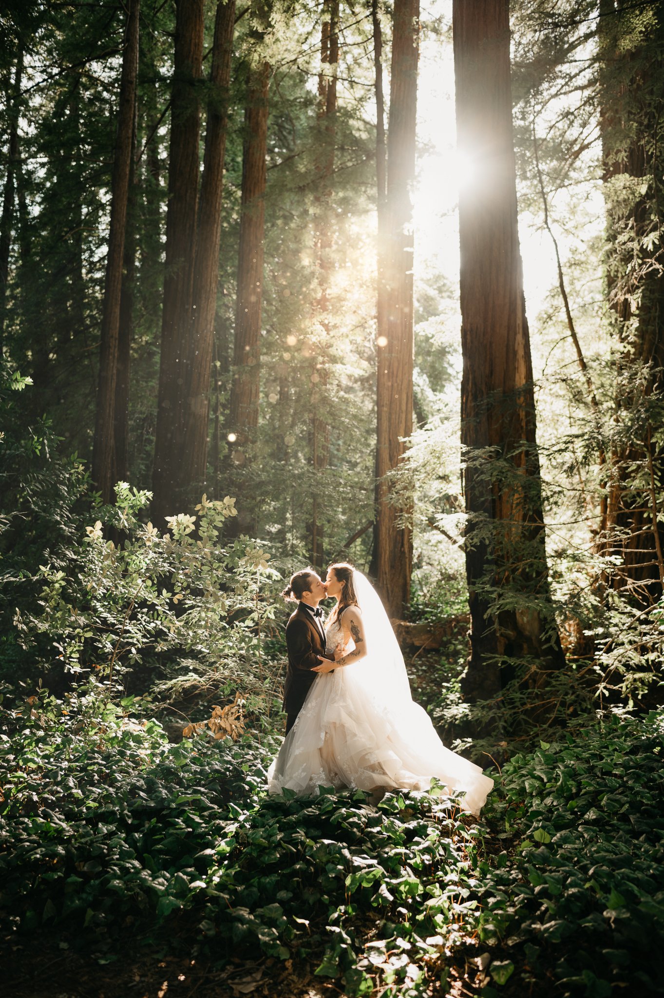 Big Sur Forest wedding bride and groom standing in forest sharing a kiss with sun peeking through the trees