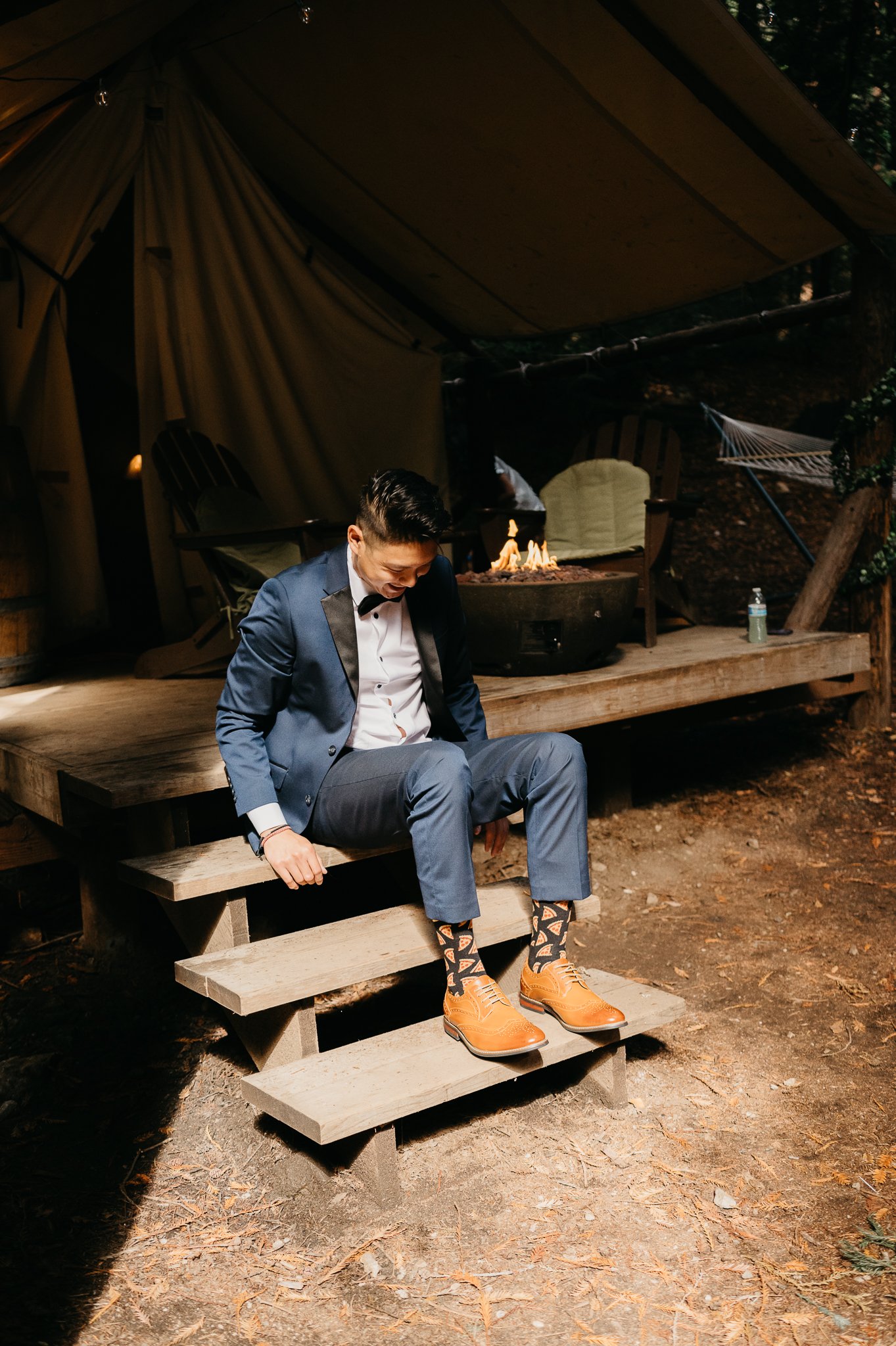 Groom getting ready for wedding ceremony at Ventana Big Sur Glamping site sitting on stairs in blue suit and tie putting on his dress shoes