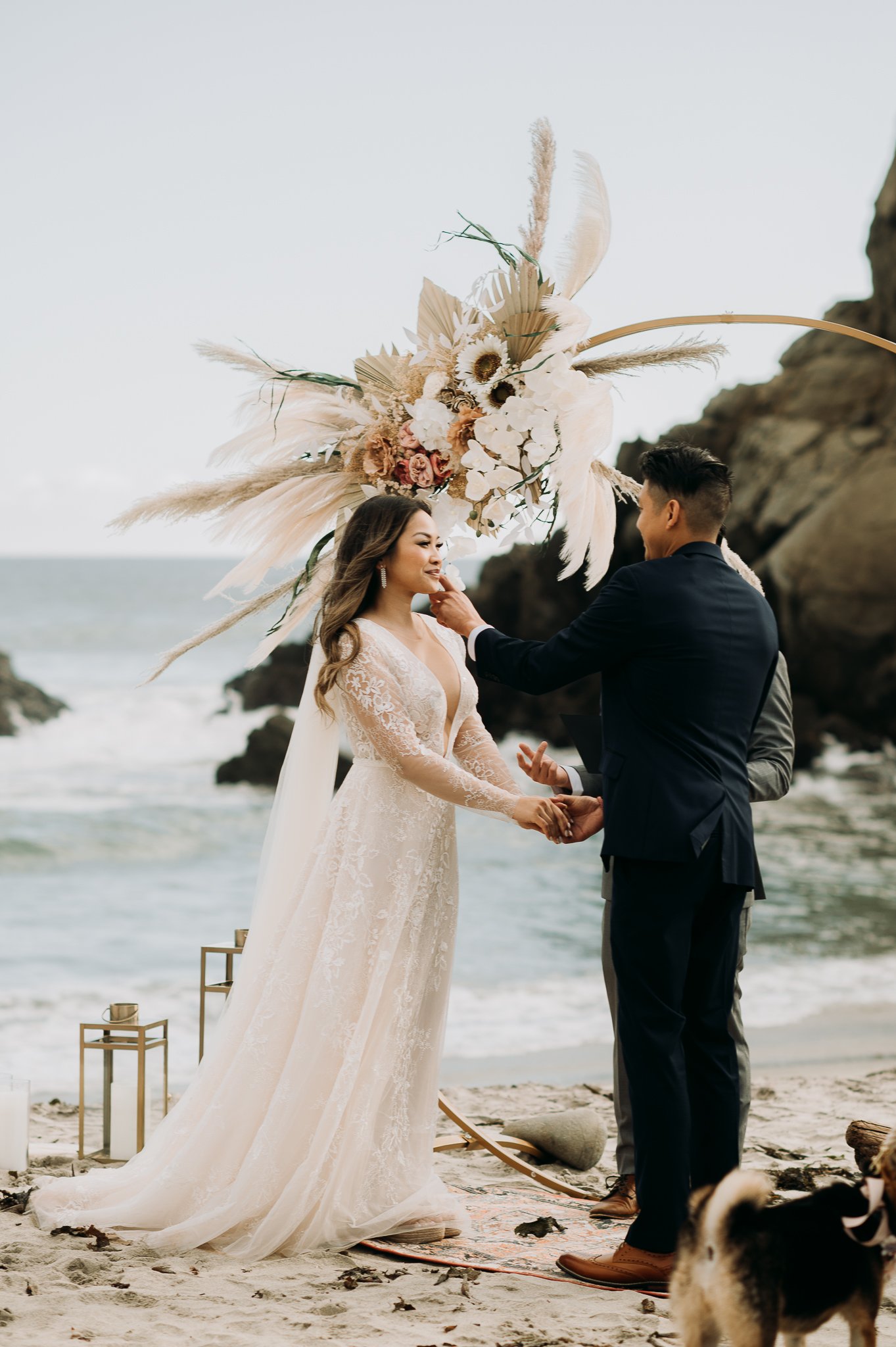 Bride and groom in wedding attire standing under a wedding arch with white flowers and feathers at Pfeiffer Beach Big Sur