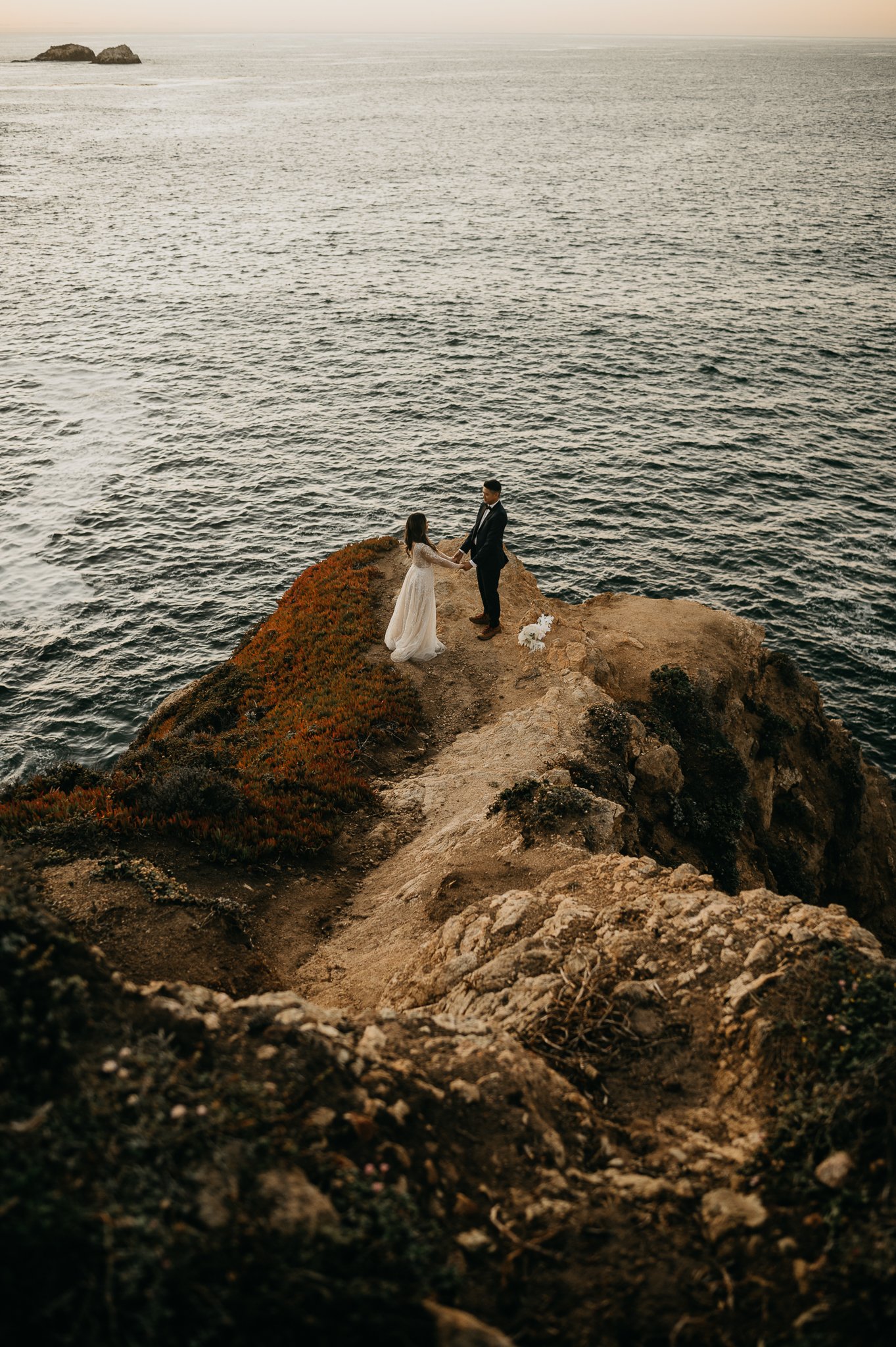 Bride and groom in wedding attire standing on a cliff with Pacific Ocean in background holding hands