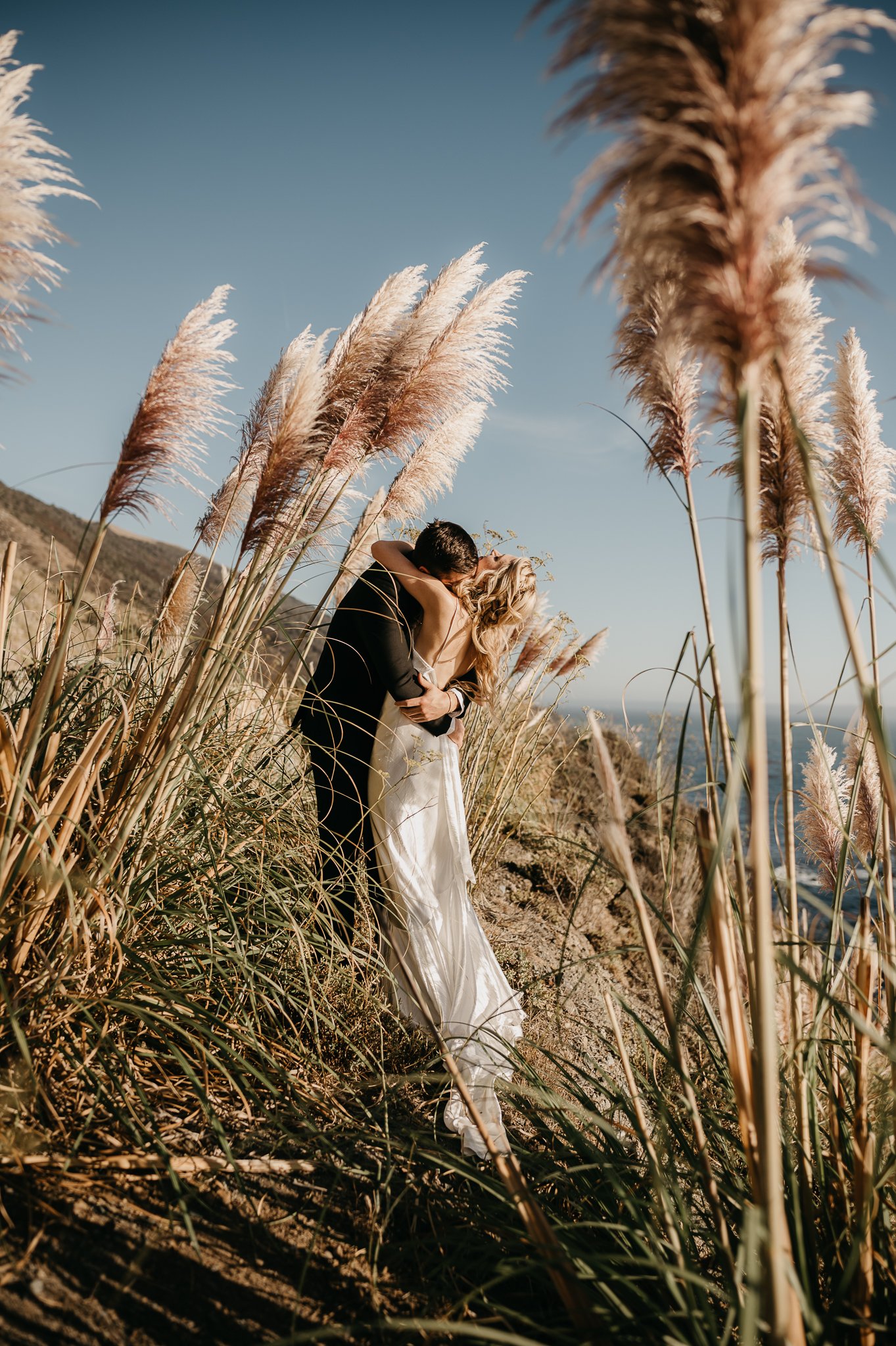 Bride and groom on Big Sur Coast in wedding attire hugging in tall grass
