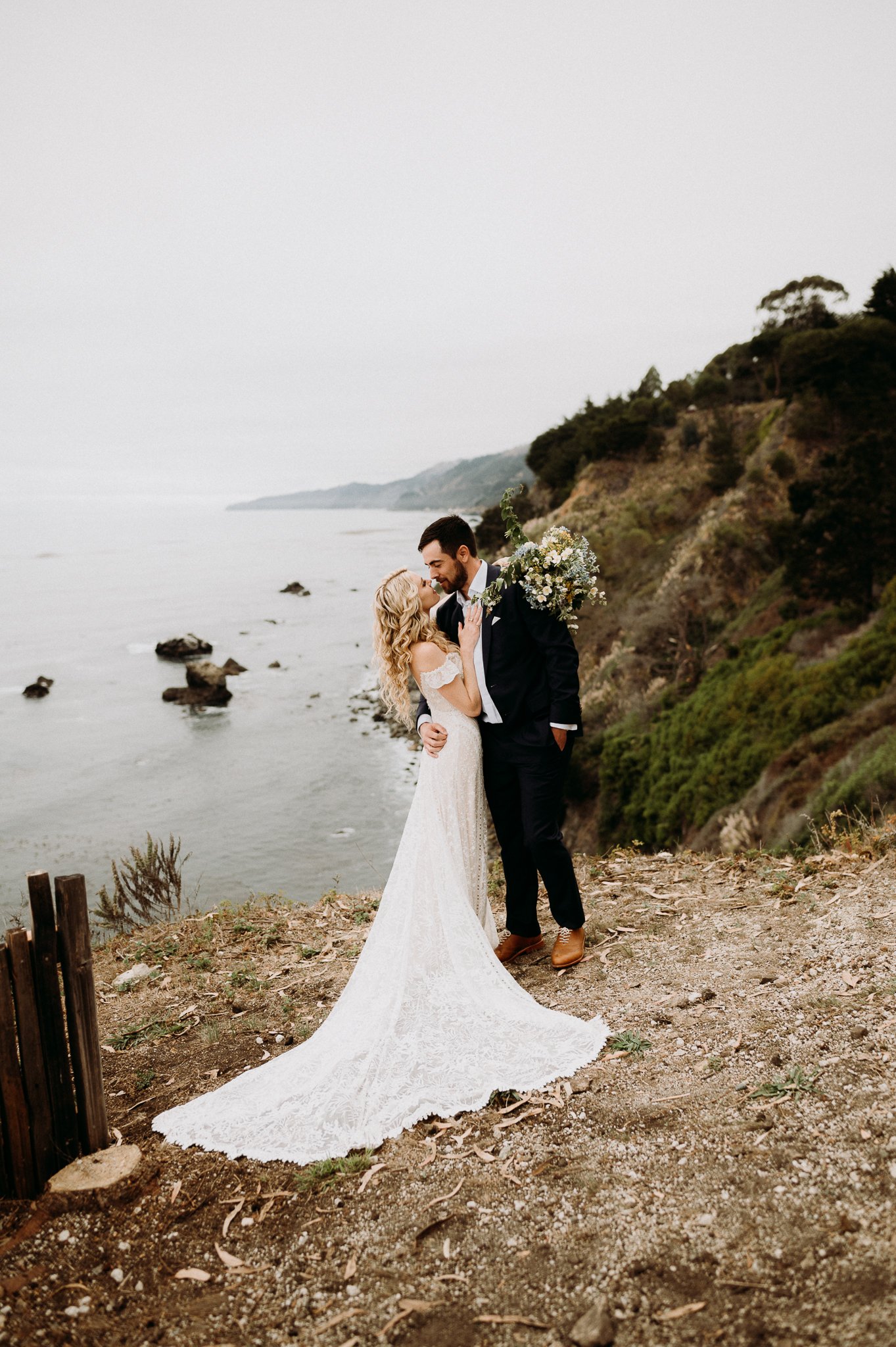 Bride and groom in wedding attire hugging on a cliff with pacific ocean in background