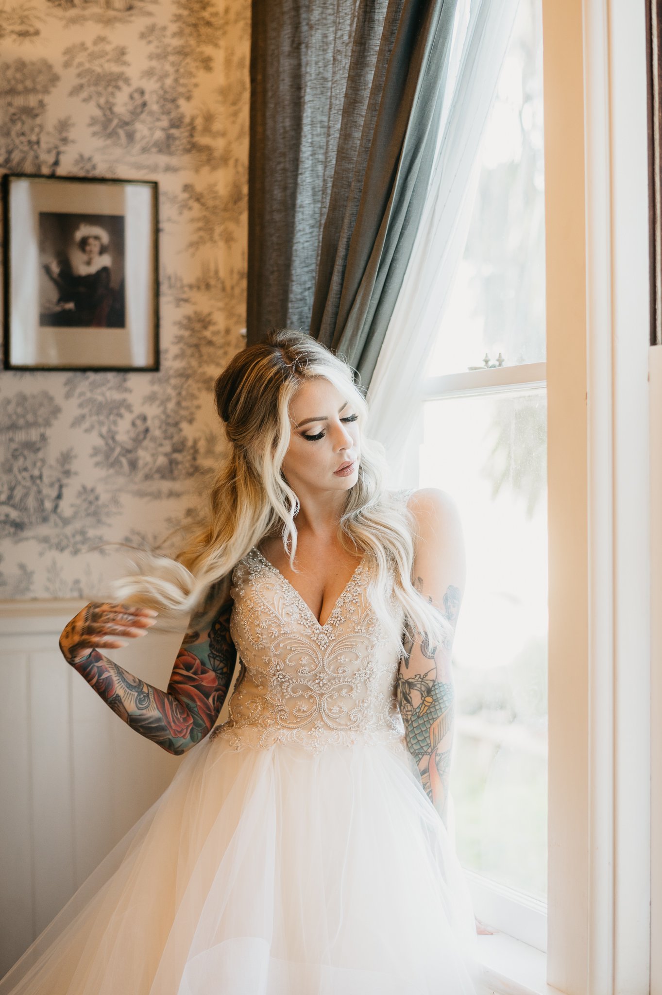 bride in wedding attire standing beside a window with sunlight shining through, she is looking down and has her fingers in the ends of her hair.