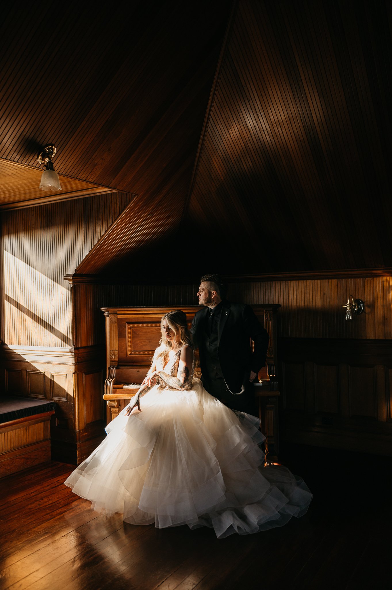 Bride and groom in wedding attire in the ballroom of wedding venue sitting on piano bench overlooking dance floor surrounded by beautiful paneled wood walls.