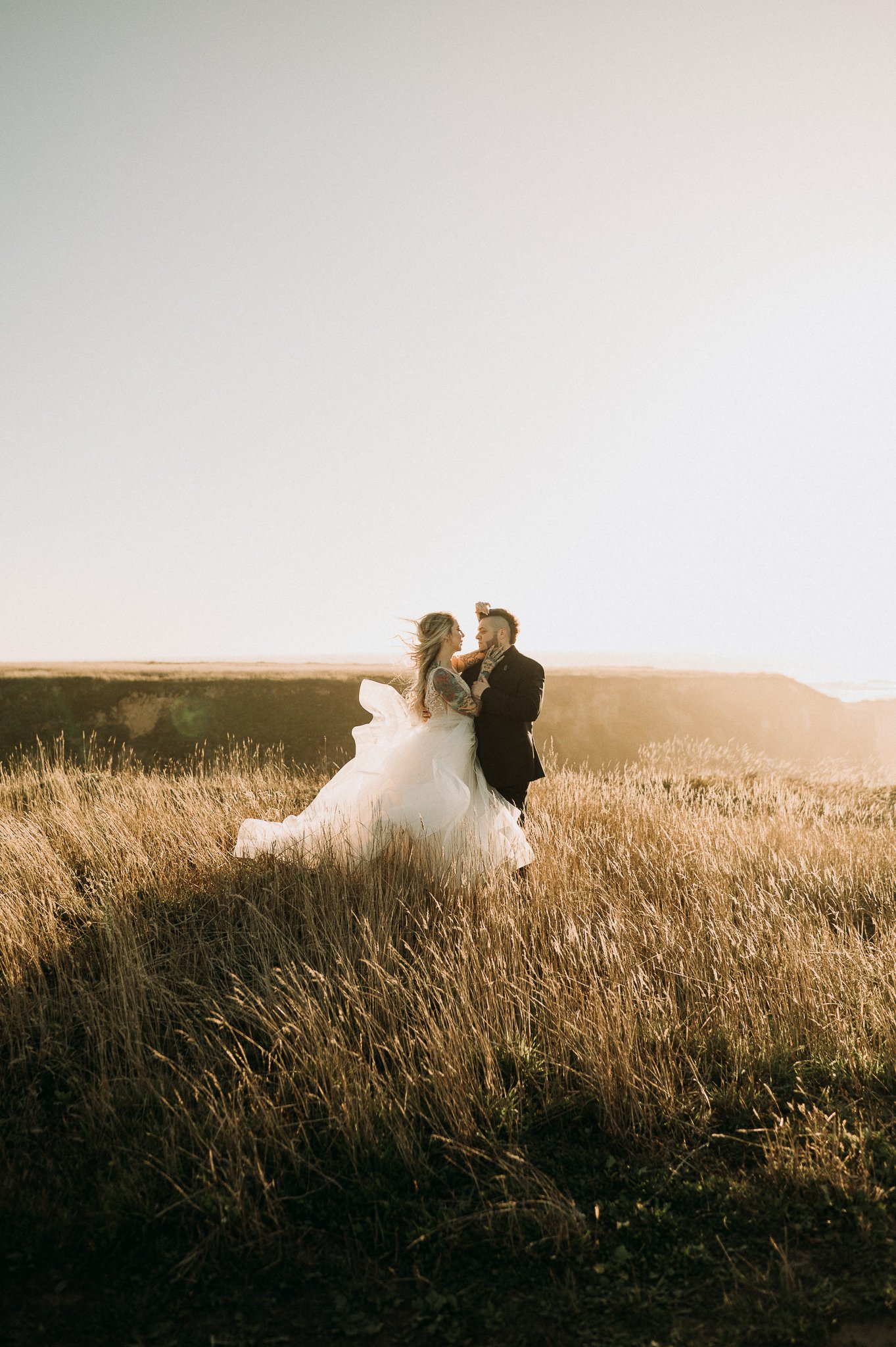 Bride and groom on open grassy field embracing each other with the wind blowing the brides gown in the wind, with the sun shining in the background.
