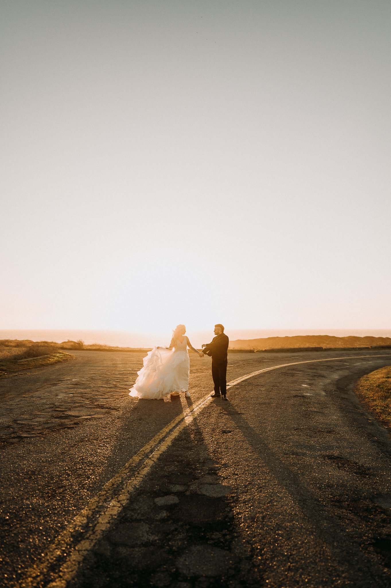 bride and groom in wedding attire on a road with yellow divider lines in center looking into each other eyes and holding hands on wedding day in Mendocino, California