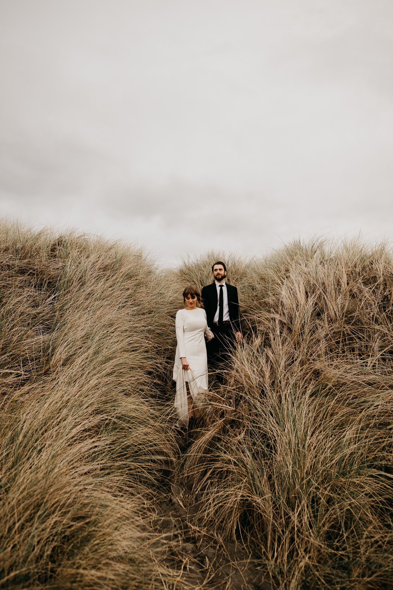 Bride and groom in wedding attire standing side by side in the tall Oregon sea grass.